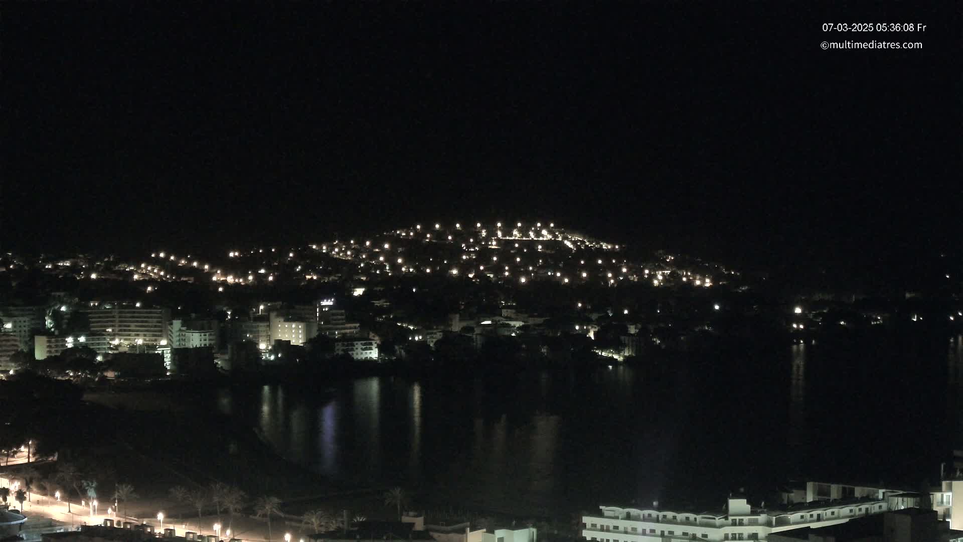A nighttime view of a city illuminated by many lights, situated beside a dark body of water reflecting the city's lights.