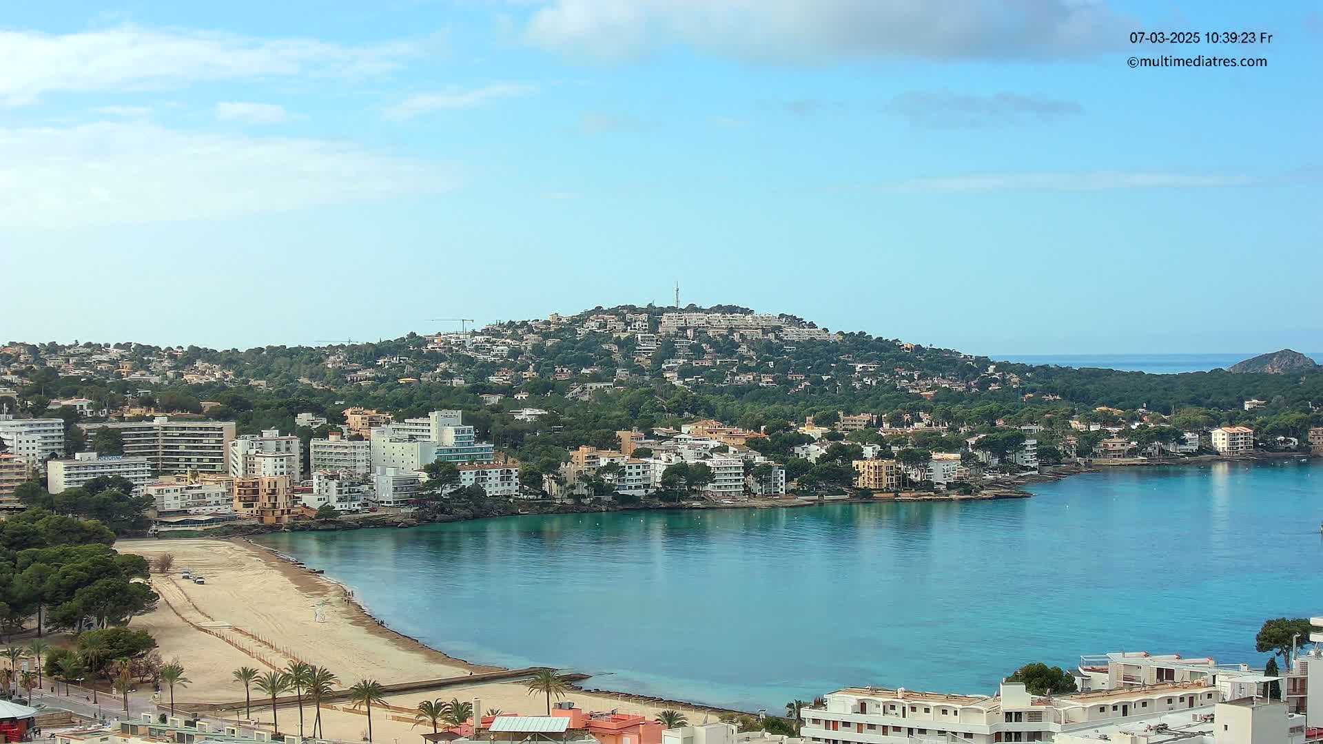 A coastal town with a sandy beach and turquoise water, nestled beneath a hill covered in buildings and greenery, under a mostly clear sky.