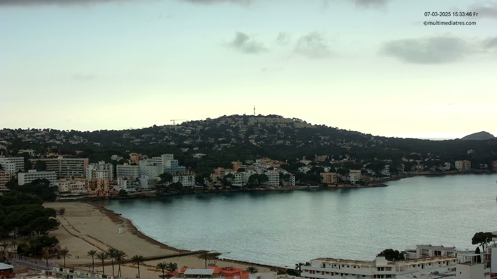 A coastal town nestled below a hill dotted with buildings, featuring a sandy beach and calm bay under a mostly cloudy sky.