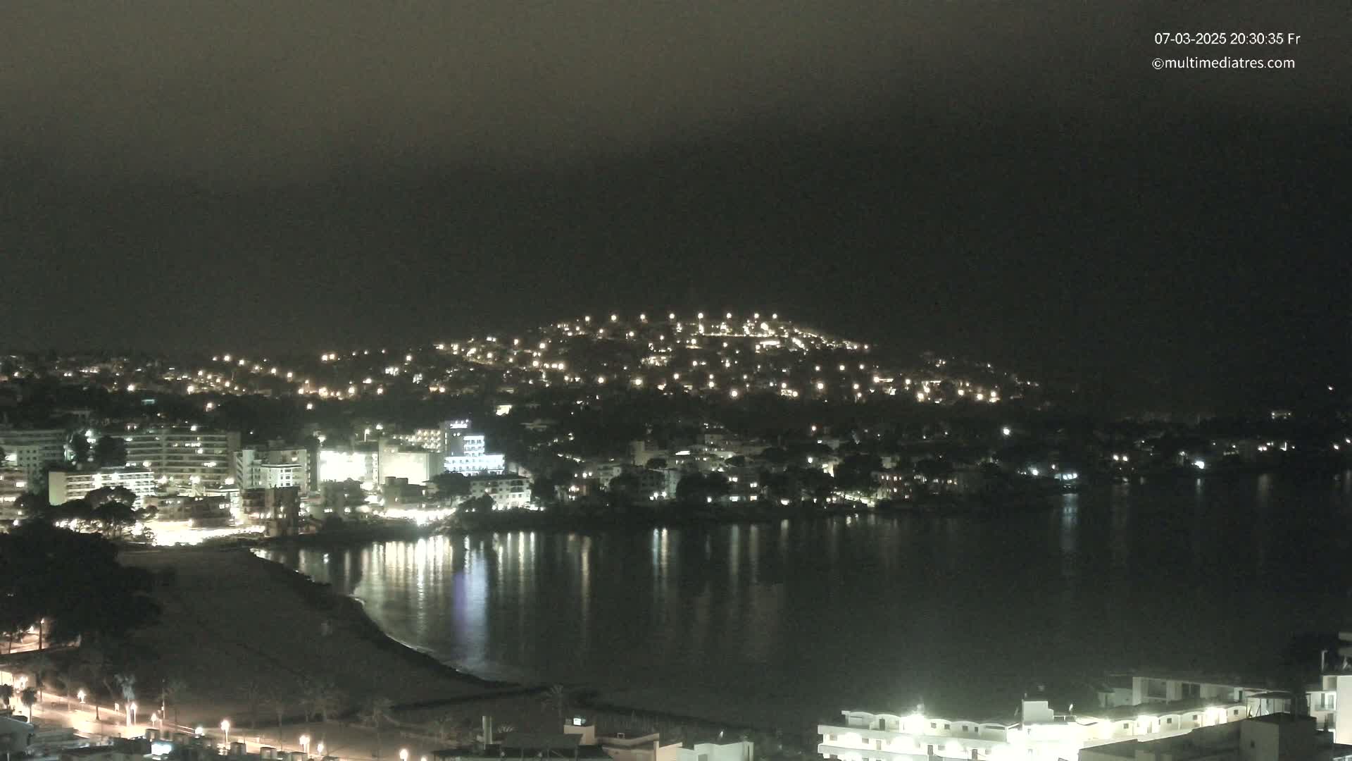A nighttime coastal town is illuminated with many lights, reflecting on a calm body of water under a dark, possibly foggy sky.