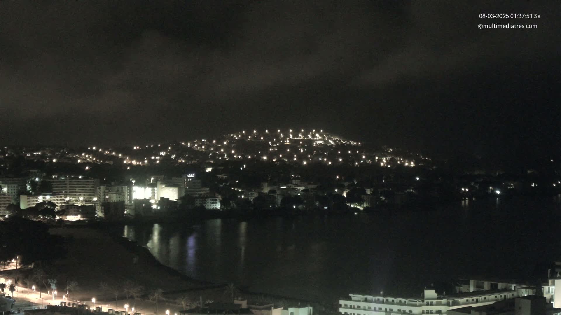 A nighttime view of a city illuminated by lights, next to a dark body of water under a cloudy sky.