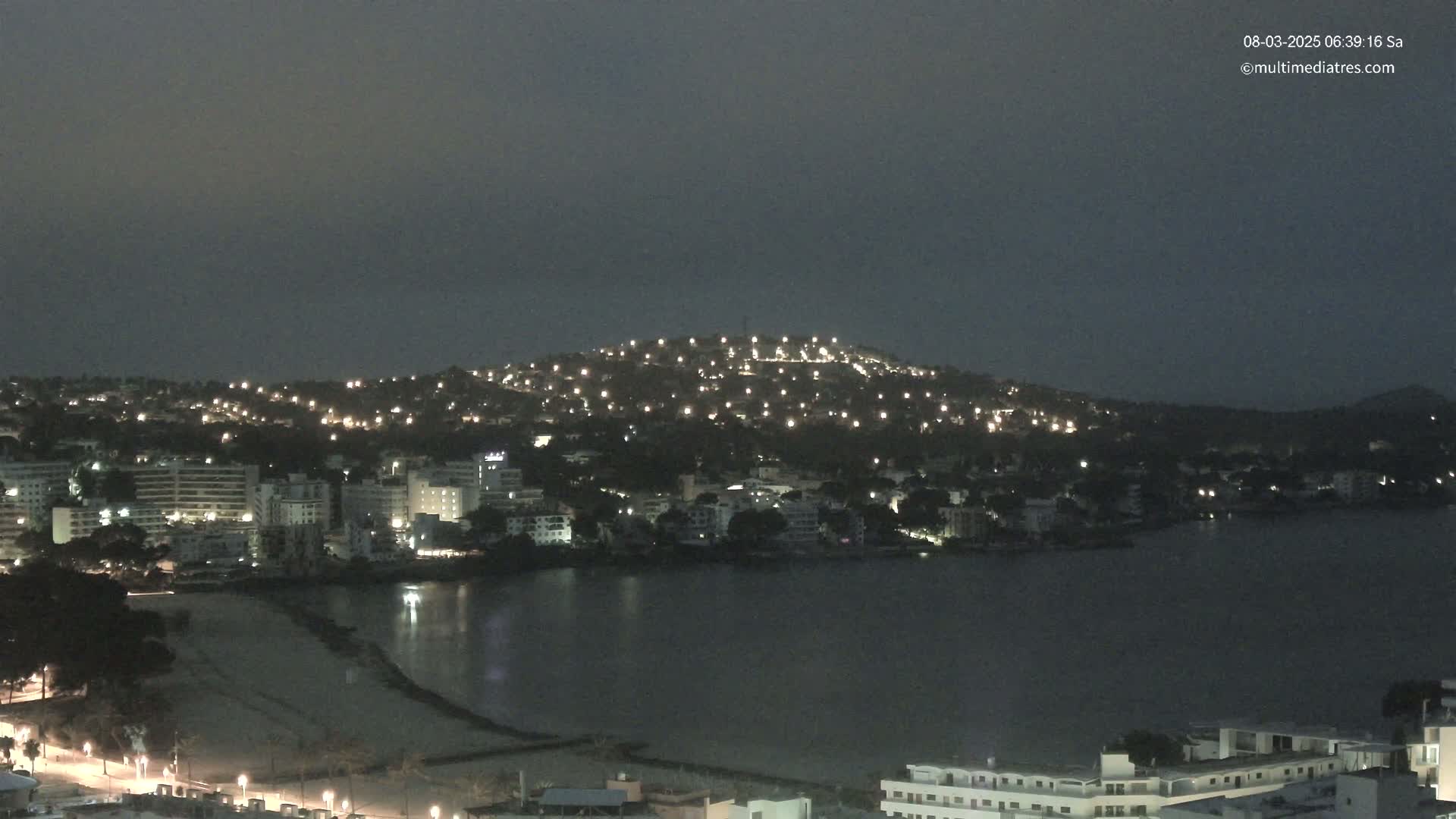 A nighttime coastal town with numerous lights illuminating buildings and a hill overlooking a calm body of water.