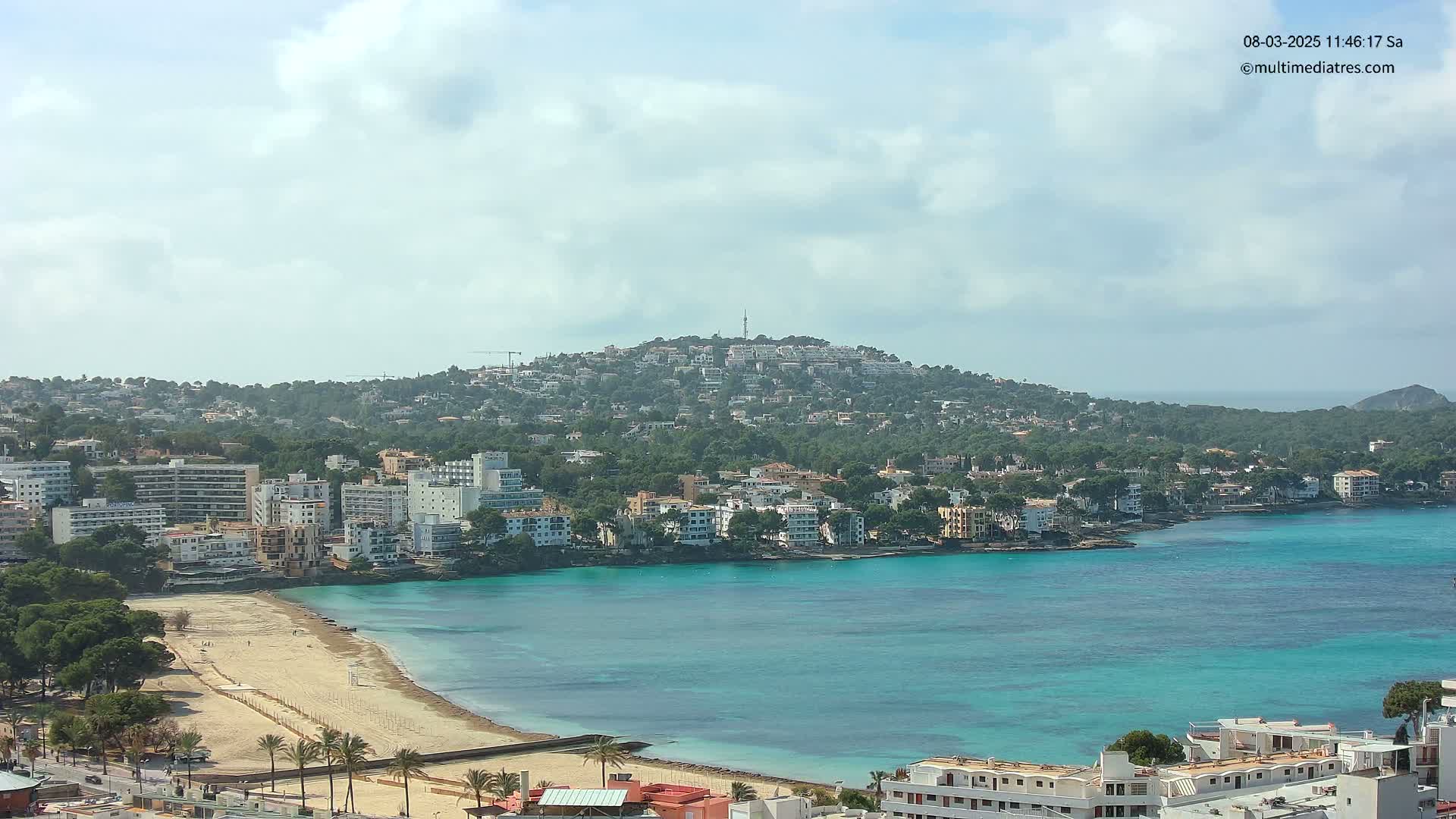 A coastal town with a sandy beach and turquoise water is nestled beneath a hill covered in buildings and vegetation under a partly cloudy sky.