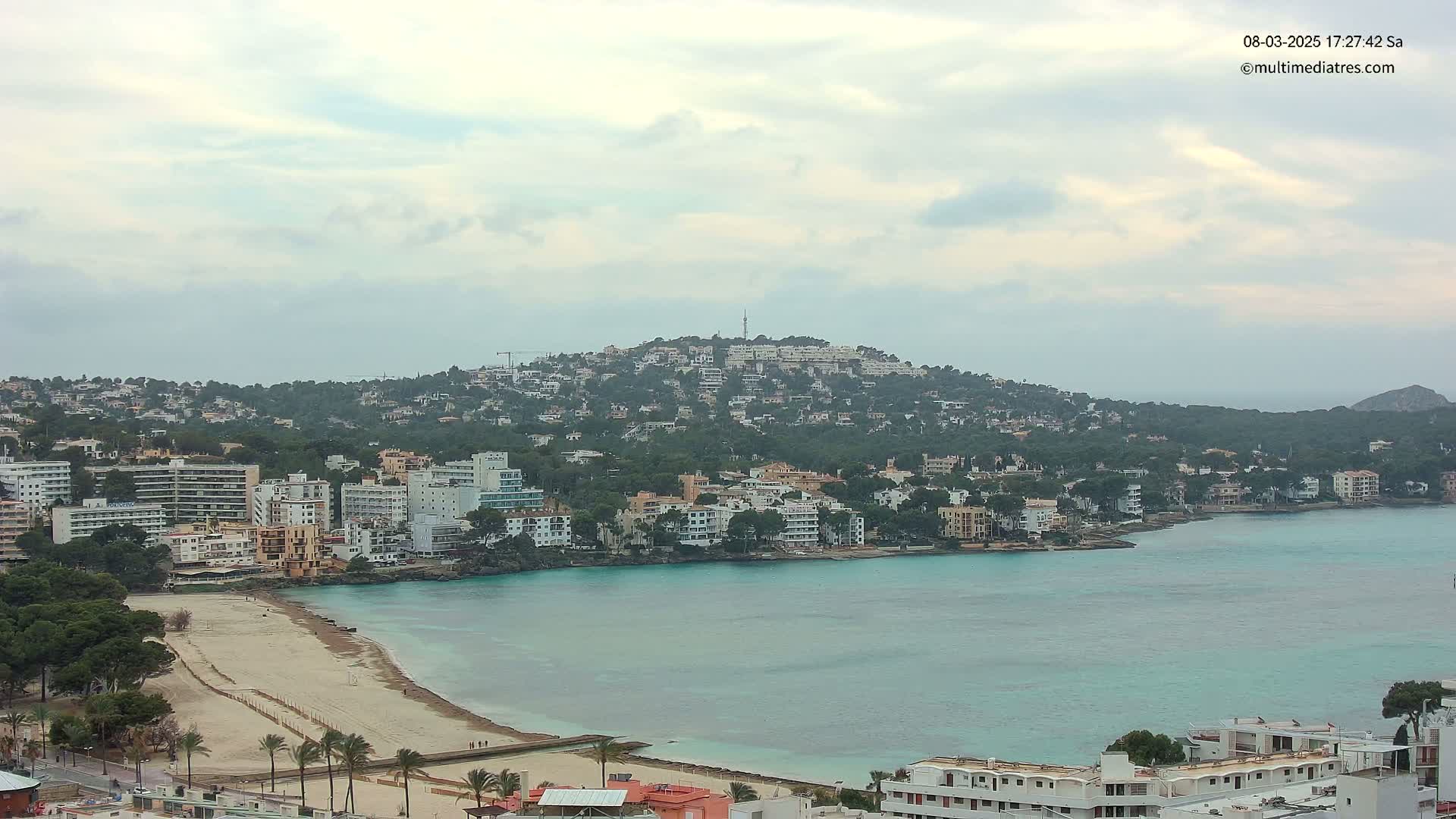 A coastal town nestled against a hill, featuring a sandy beach and calm, turquoise water under an overcast sky.