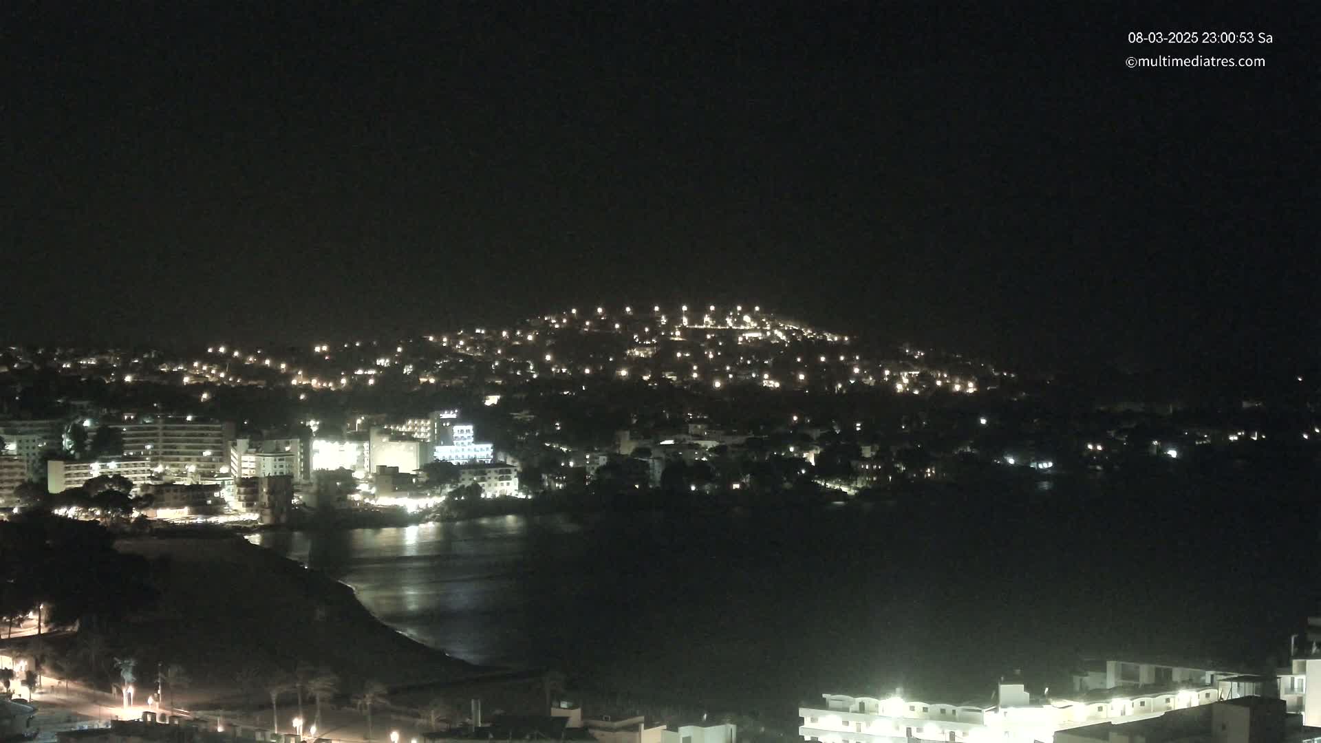 A nighttime view of a coastal town illuminated by many lights, with a dark, calm body of water in the foreground and a hilltop in the background under a clear, dark sky.