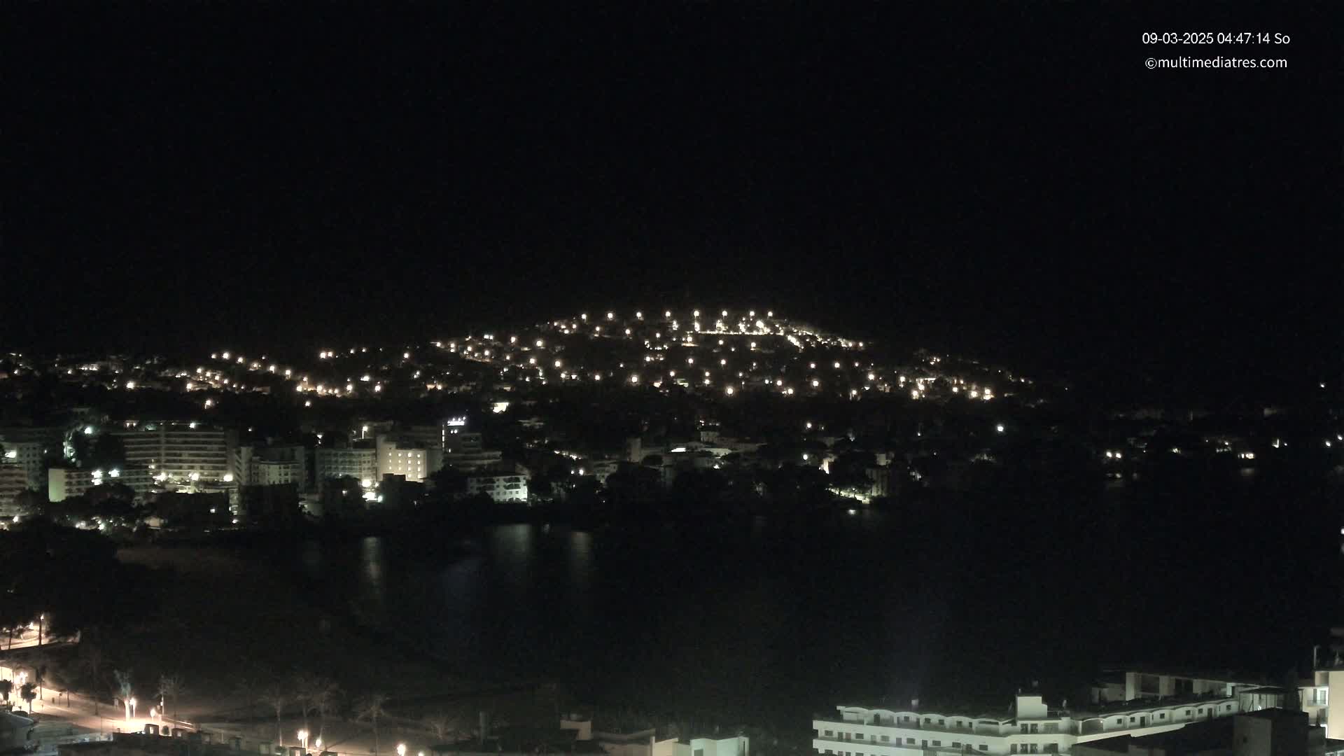 A nighttime view of a city illuminated by scattered lights, situated near a dark body of water, under a clear, dark sky.