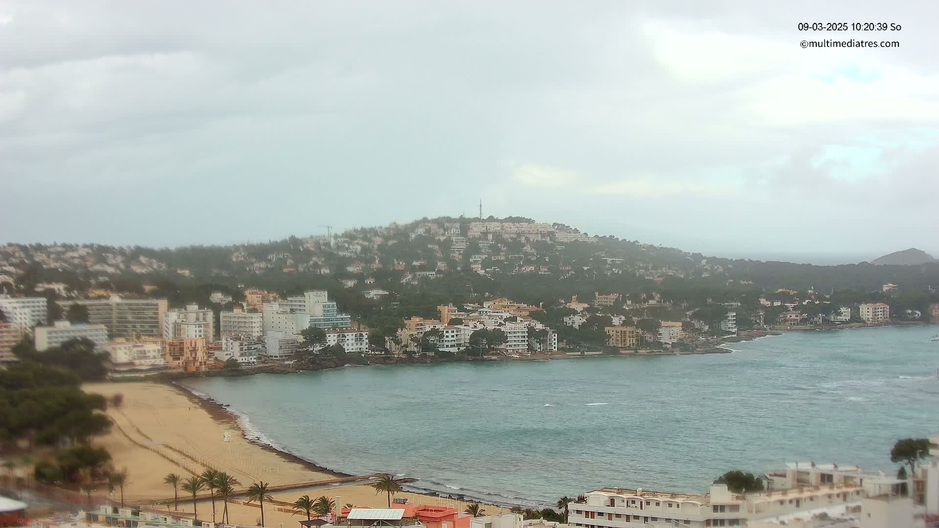 A coastal town nestled against a hill, with a sandy beach and calm, overcast water.
