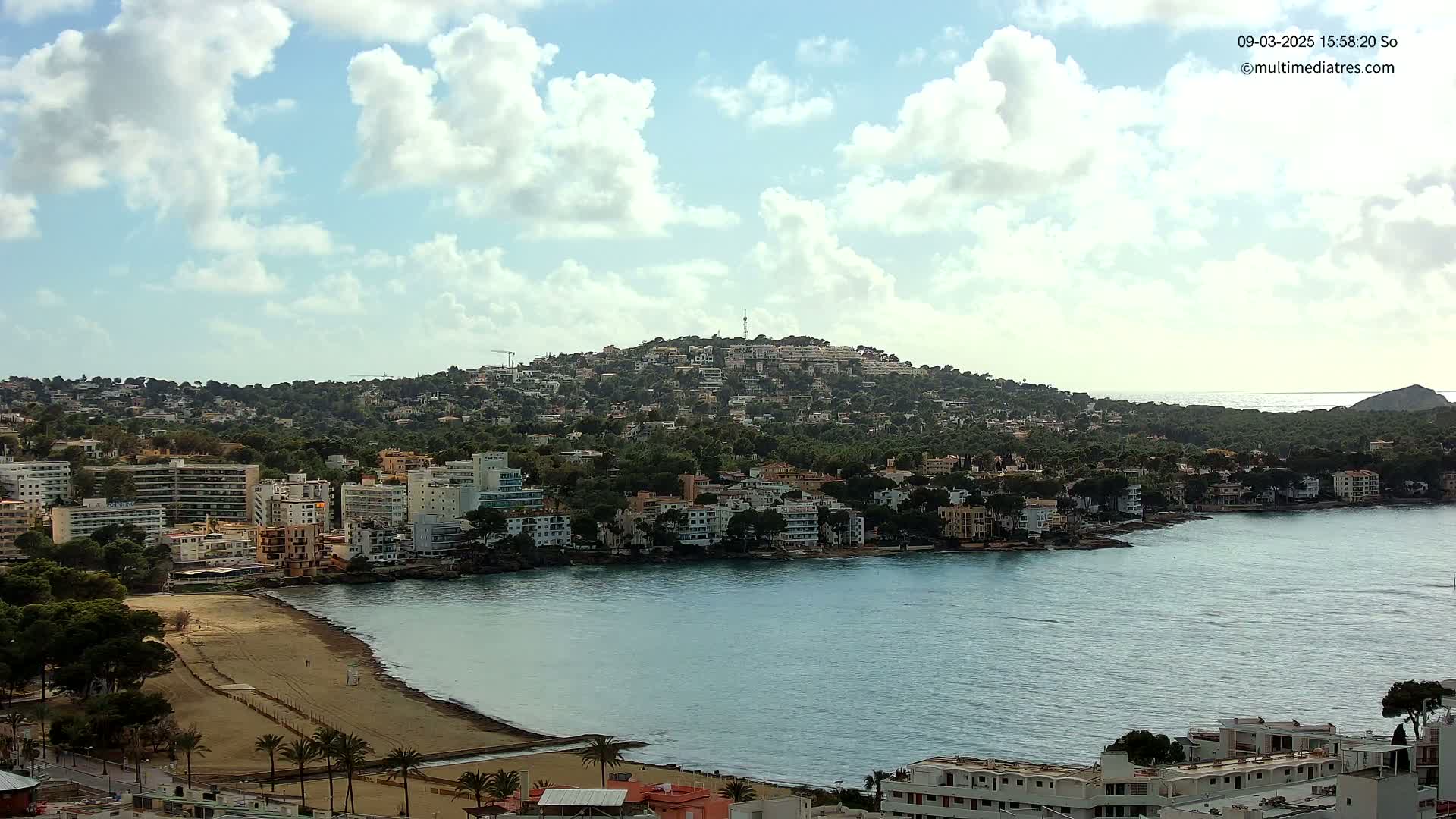 A coastal town with buildings on a hillside and along a sandy beach, overlooks a calm bay under a partly cloudy sky.