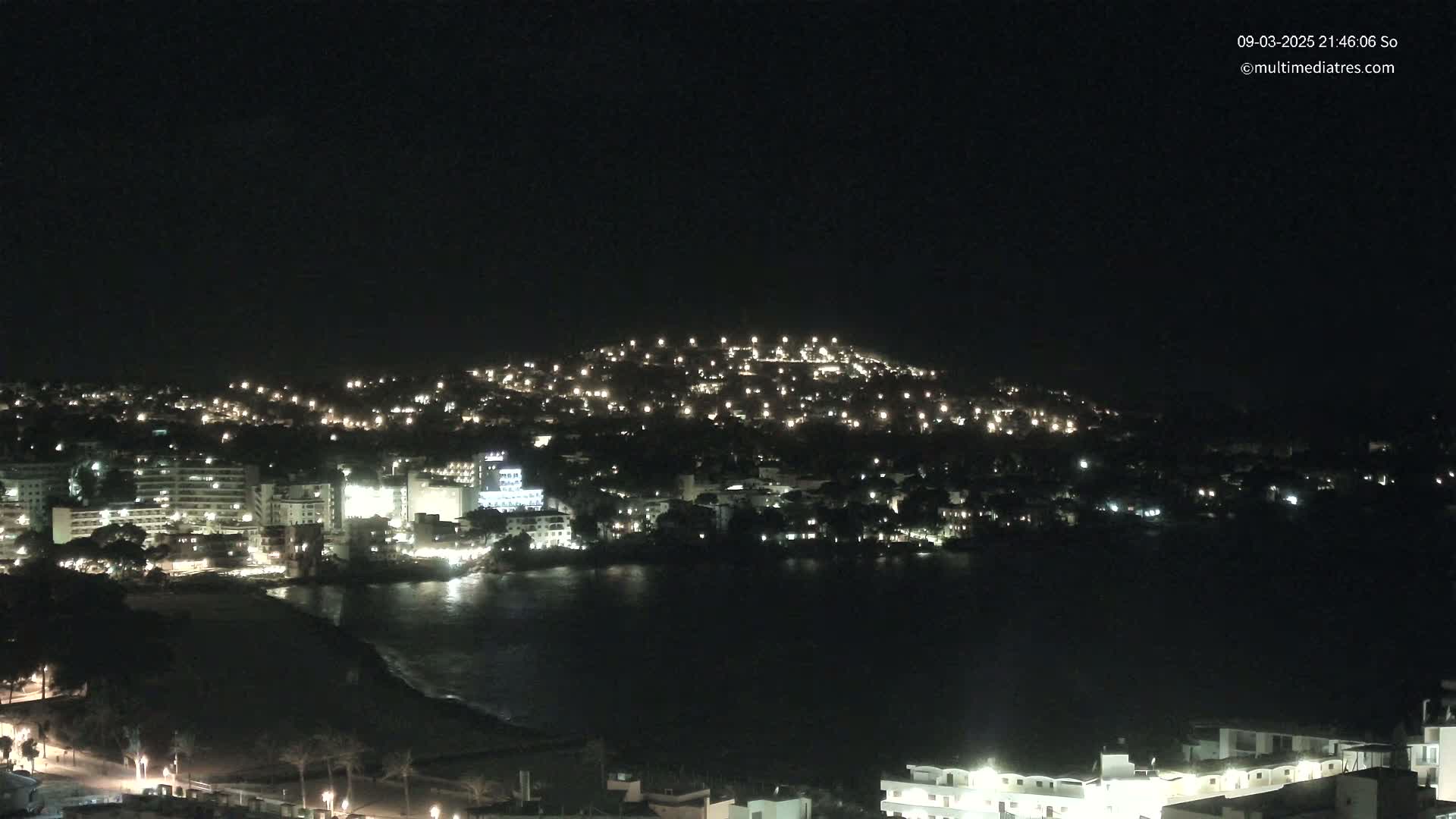 A nighttime coastal town with numerous lights illuminating buildings and streets along a dark body of water under a clear, dark sky.