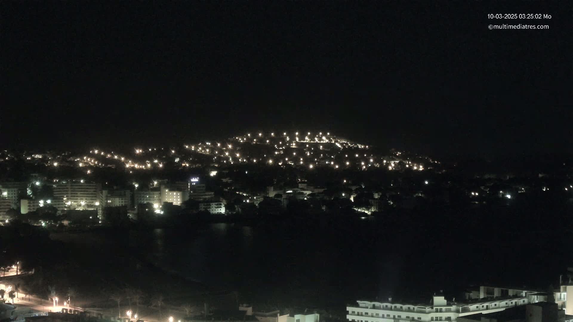 A nighttime view of a coastal town illuminated by many lights, with a hilltop also lit up in the background under a clear, dark sky.