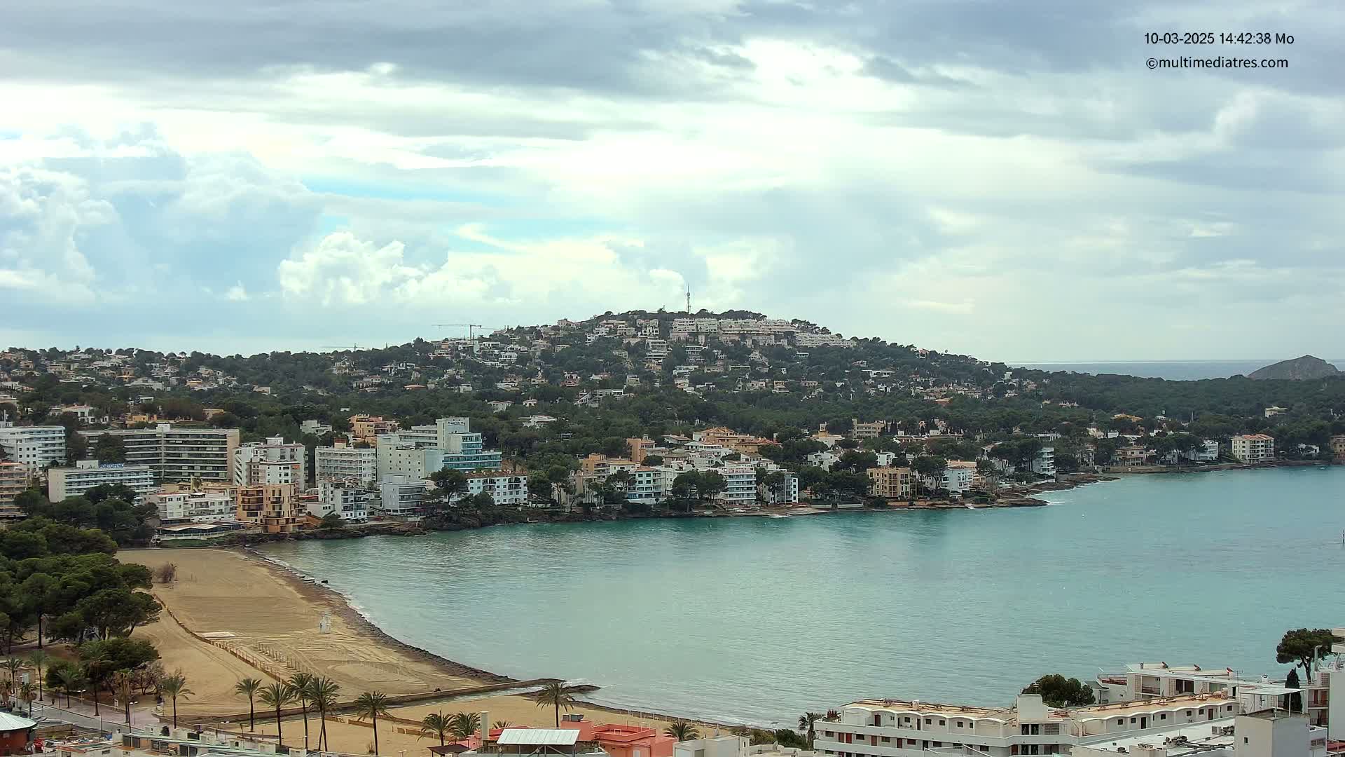 A coastal town nestled against a hill under a partly cloudy sky features a sandy beach and calm blue-green water.