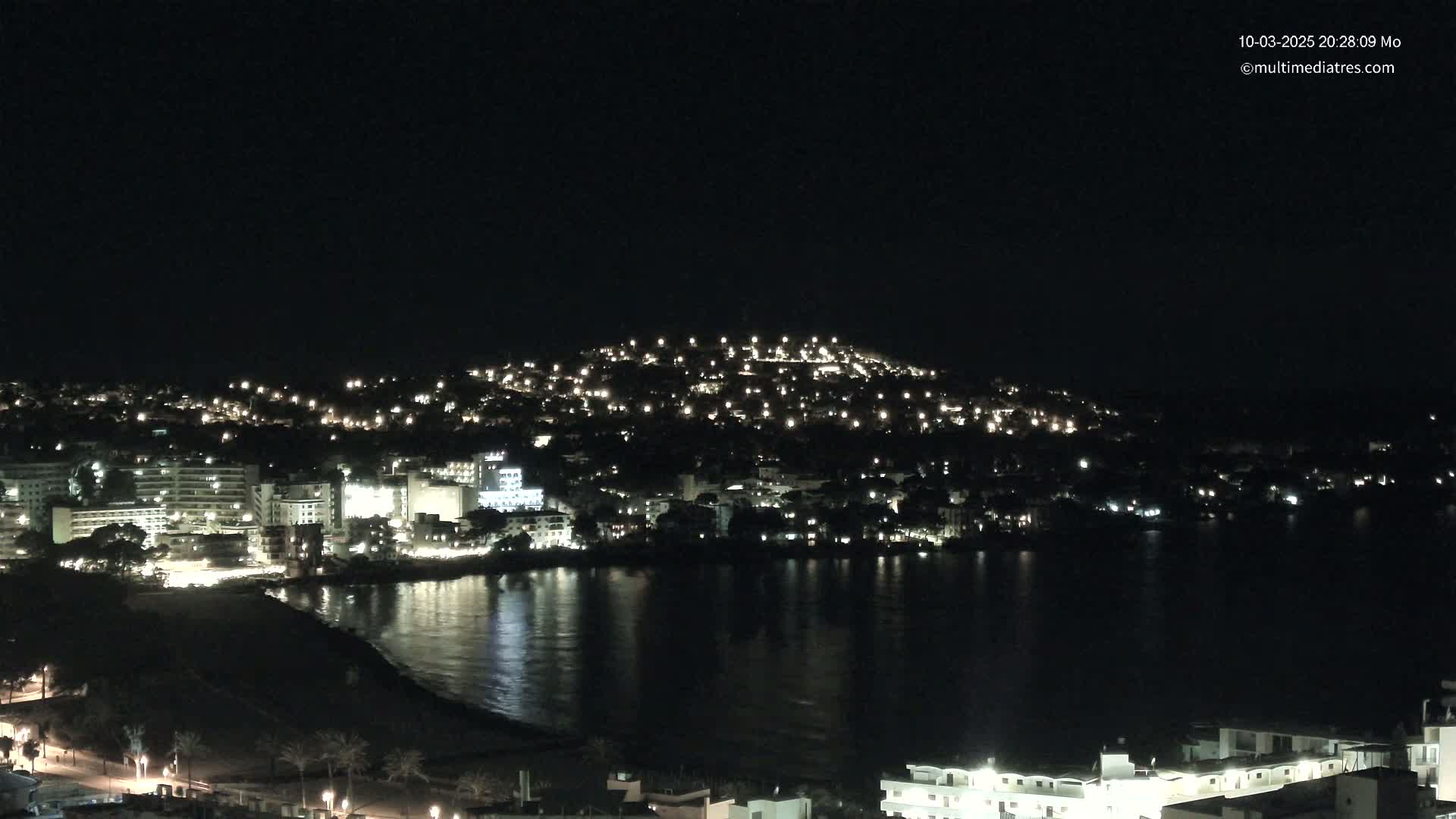A coastal town at night, illuminated by numerous lights, is reflected in a calm body of water under a clear, dark sky.