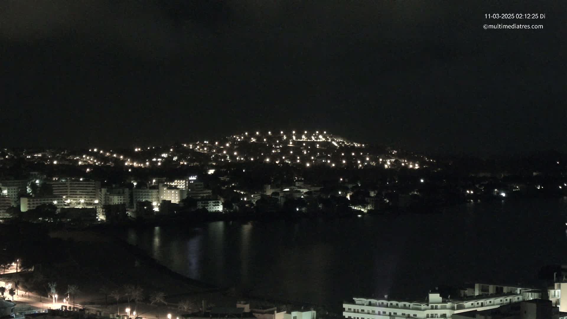 A nighttime view shows a city illuminated by many lights, situated on a hill next to a dark body of water under a dark, clear sky.