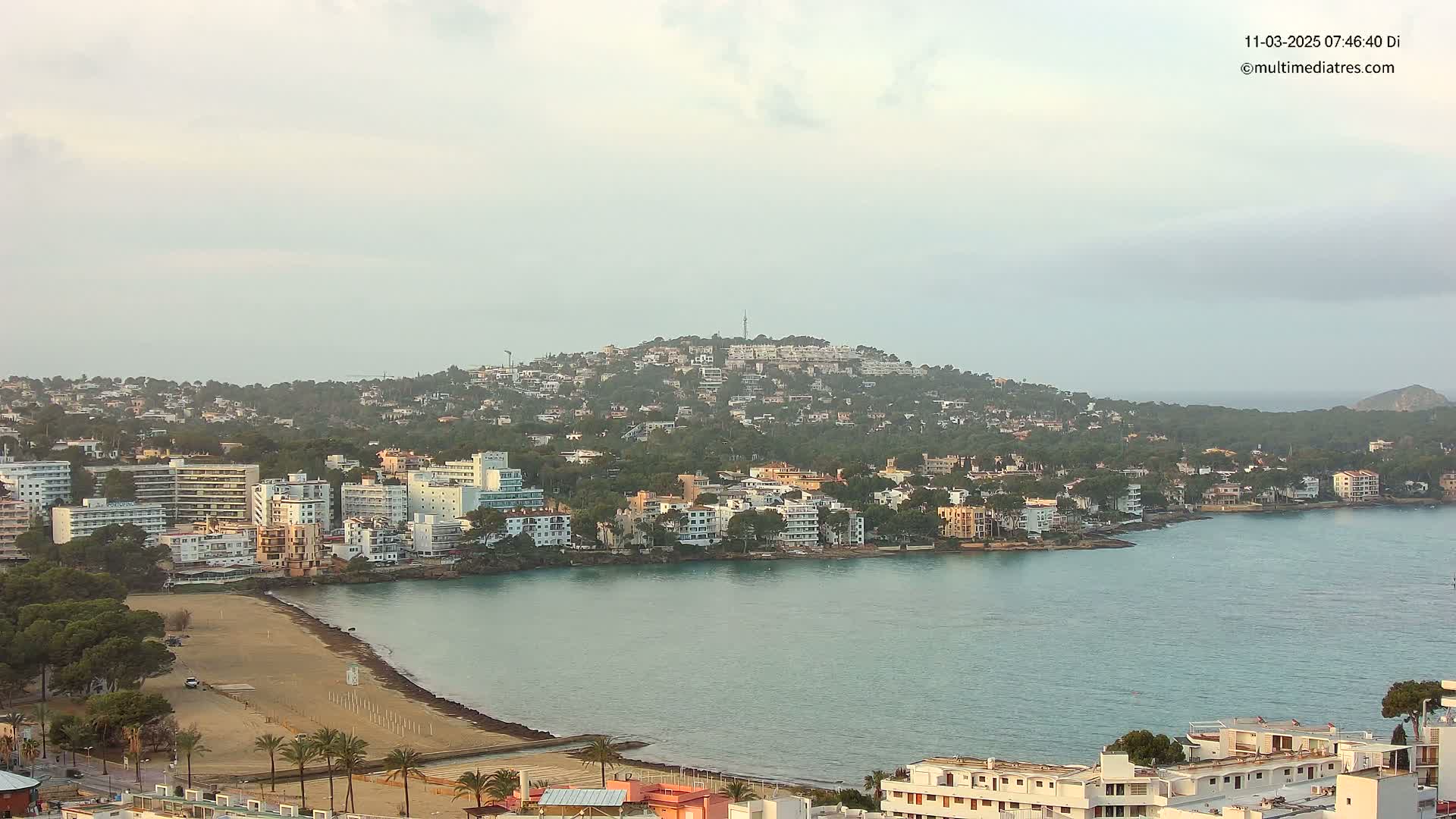 A coastal town with a sandy beach and many buildings nestled at the base of a hill, under a mostly cloudy sky.
