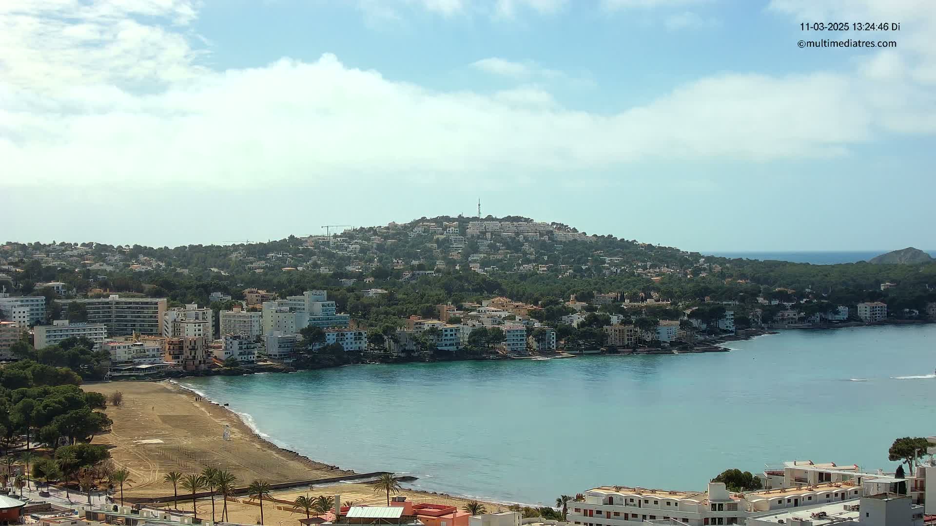 A coastal town with a sandy beach and calm turquoise water is nestled at the base of a hill covered in buildings and trees under a partly cloudy sky.