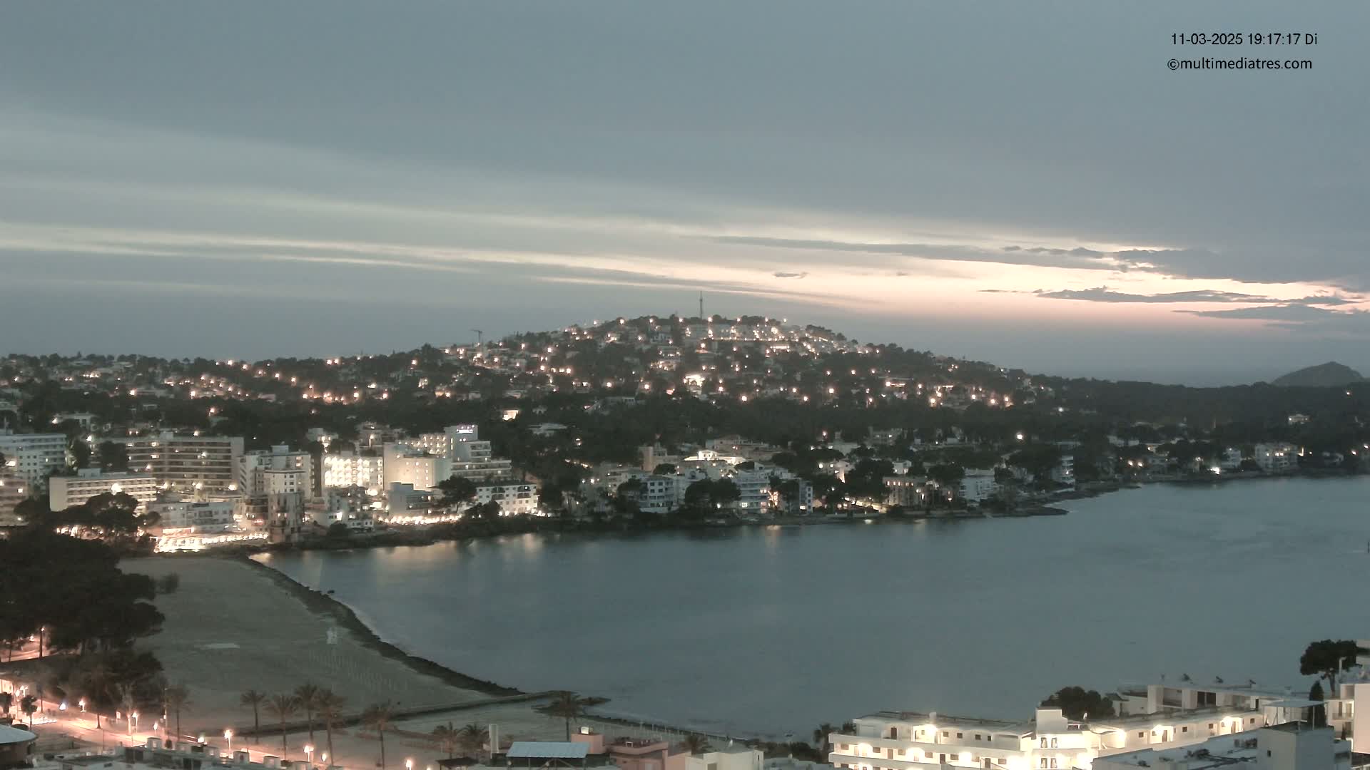A coastal town at dusk, with its lights illuminated against a calm bay under a mostly cloudy sky.