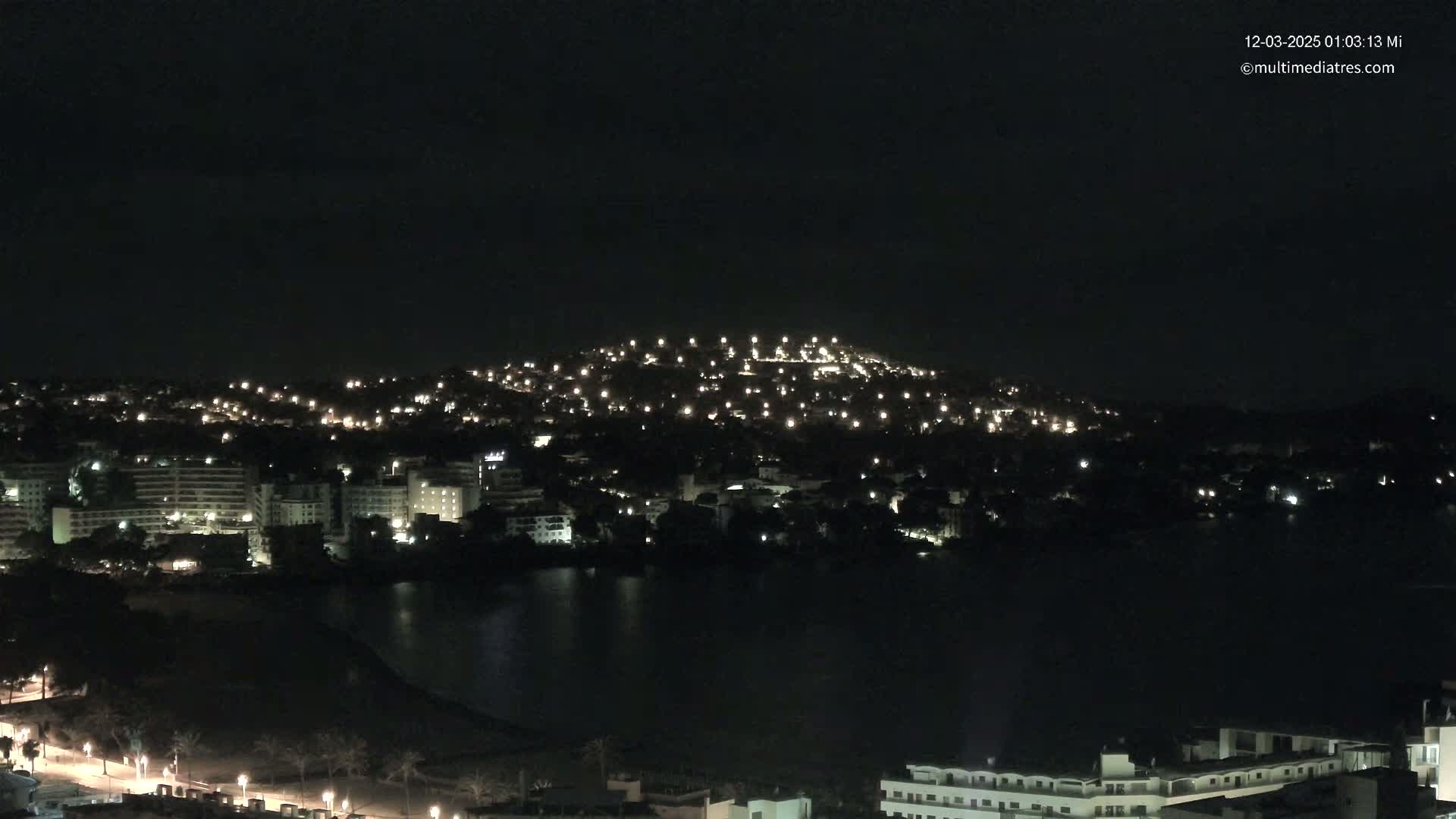 A nighttime view of a city illuminated by lights, nestled beside a dark body of water, under a clear, dark sky.