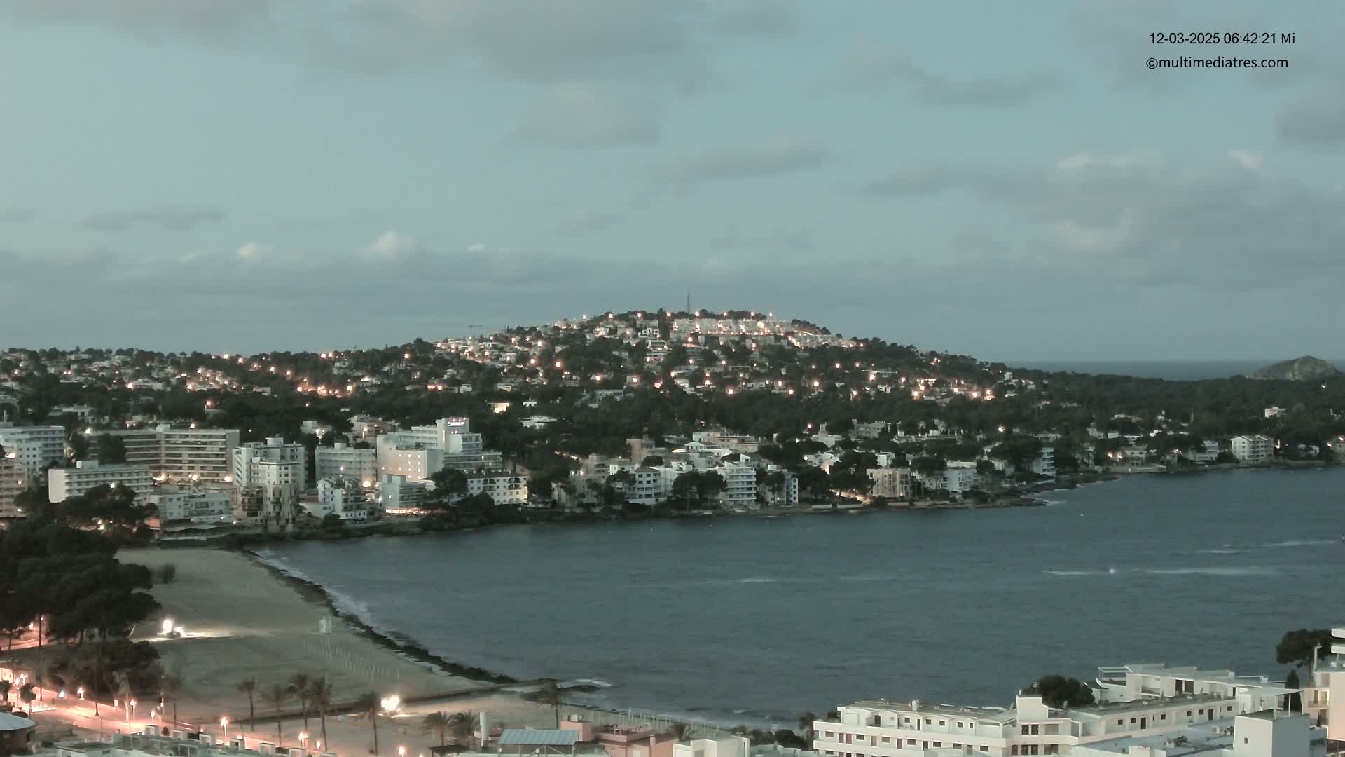 A coastal town at twilight, with lights illuminating buildings on a hill overlooking a calm bay and sandy beach under a mostly cloudy sky.