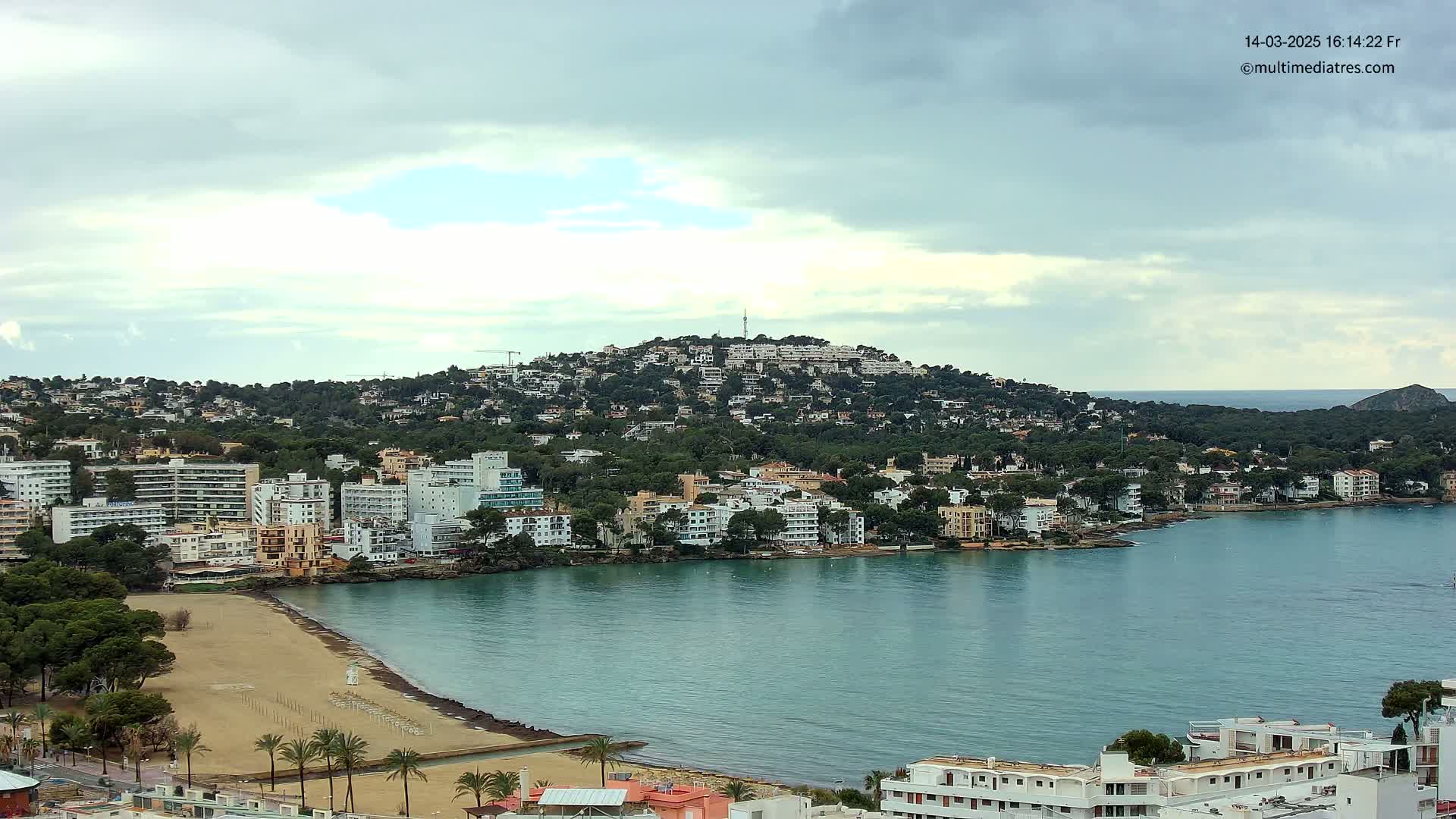 A coastal town with sandy beach and numerous buildings sprawls across a hillside overlooking a calm bay under a cloudy sky.