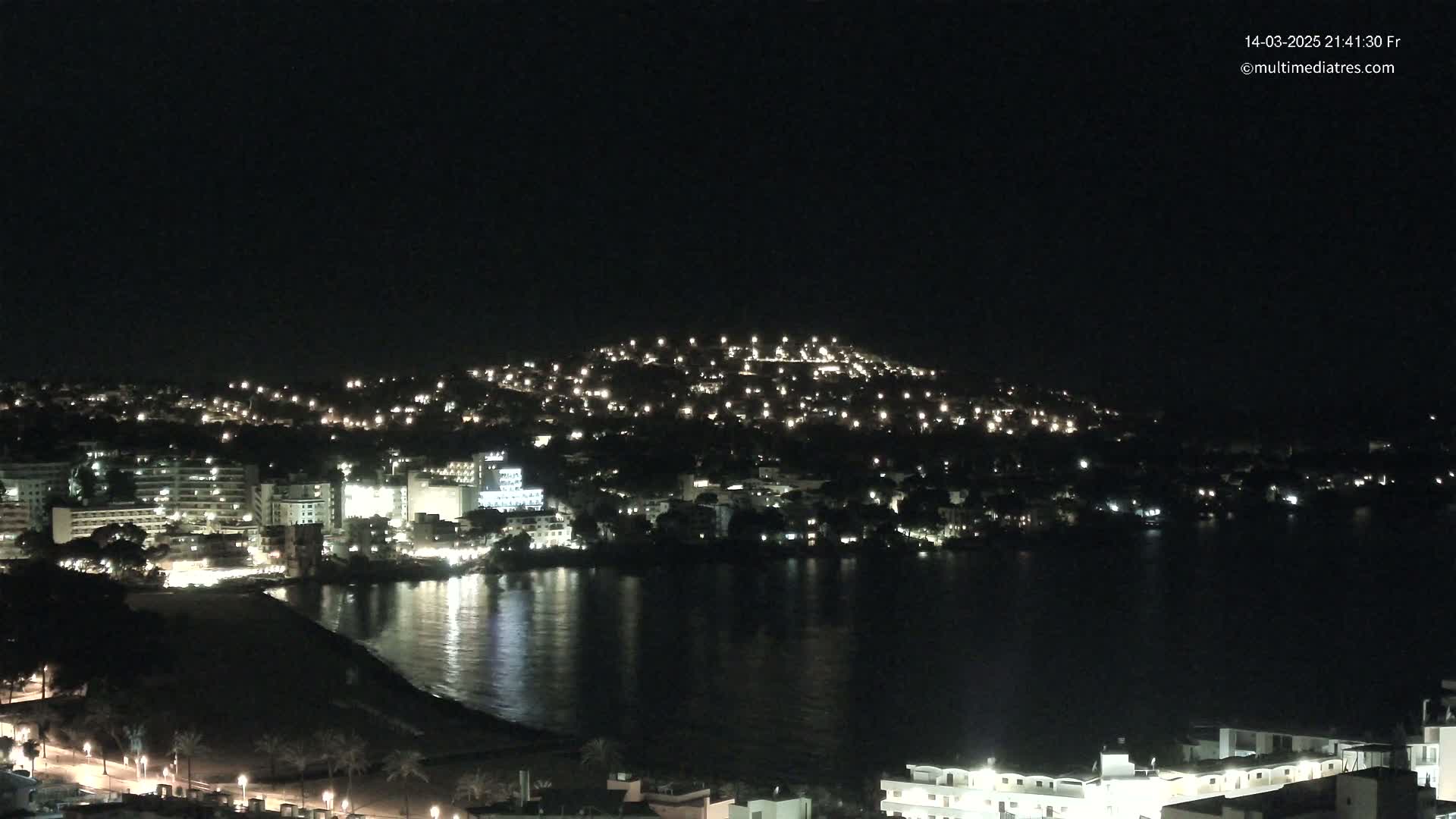 A nighttime coastal city is illuminated by countless lights, nestled against a dark hill, with a calm body of water reflecting the city lights.