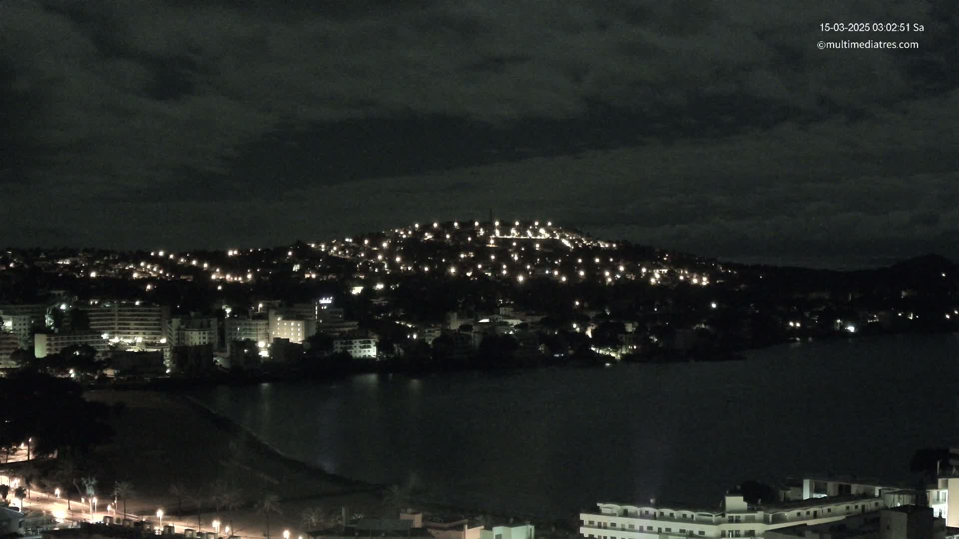 A nighttime view of a city and hill illuminated by numerous lights, beside a dark body of water under a cloudy sky.