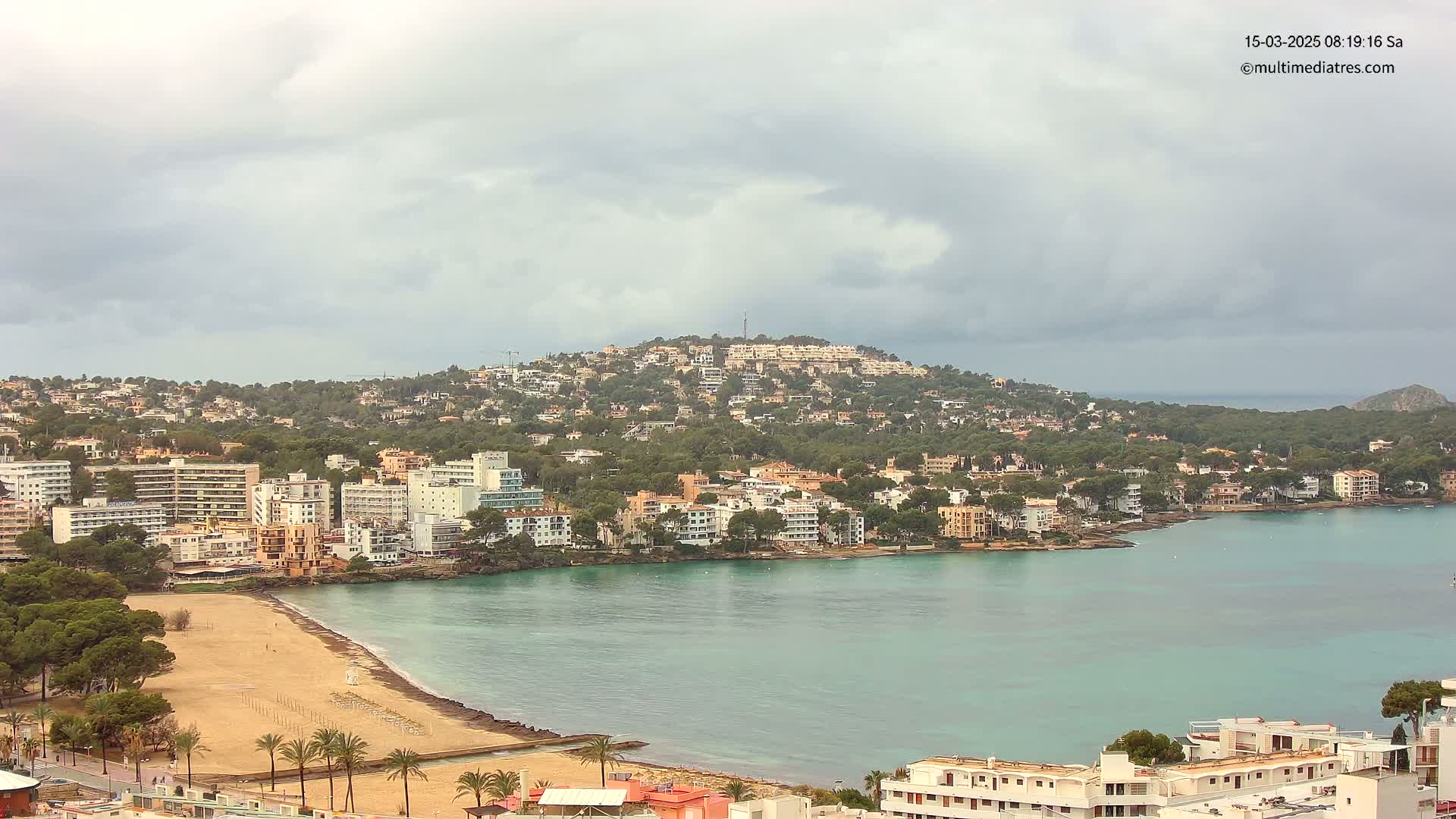 A coastal town with a sandy beach and turquoise water sits beneath a hill covered in buildings and trees under an overcast sky.
