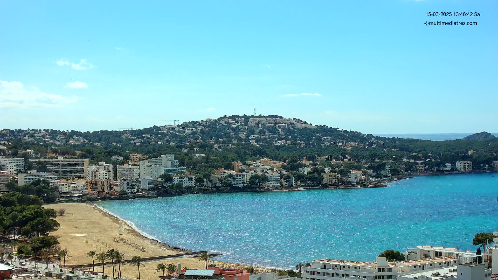 A coastal town with sandy beach and turquoise water sits below a hill covered in buildings and greenery under a clear blue sky.