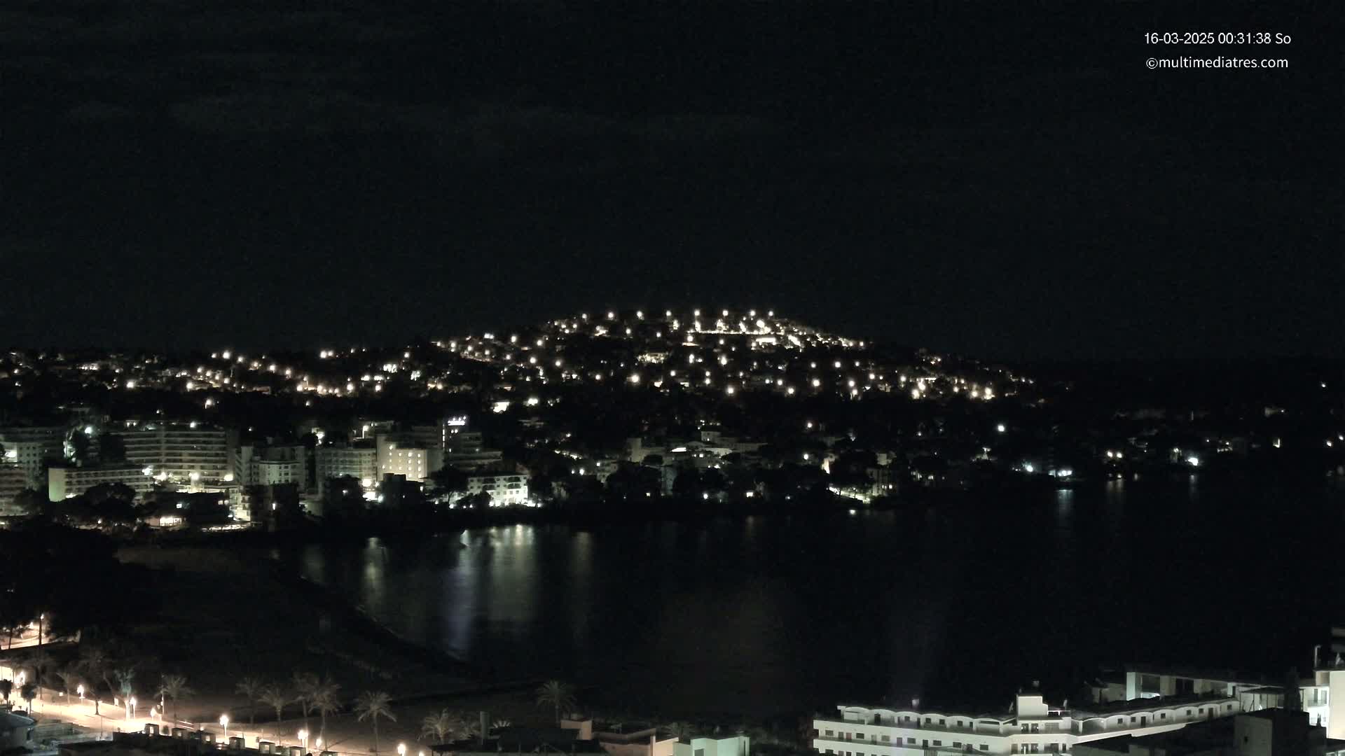 A nighttime view of a city sprawling across a hill and down to a dark body of water, under a clear, dark sky.