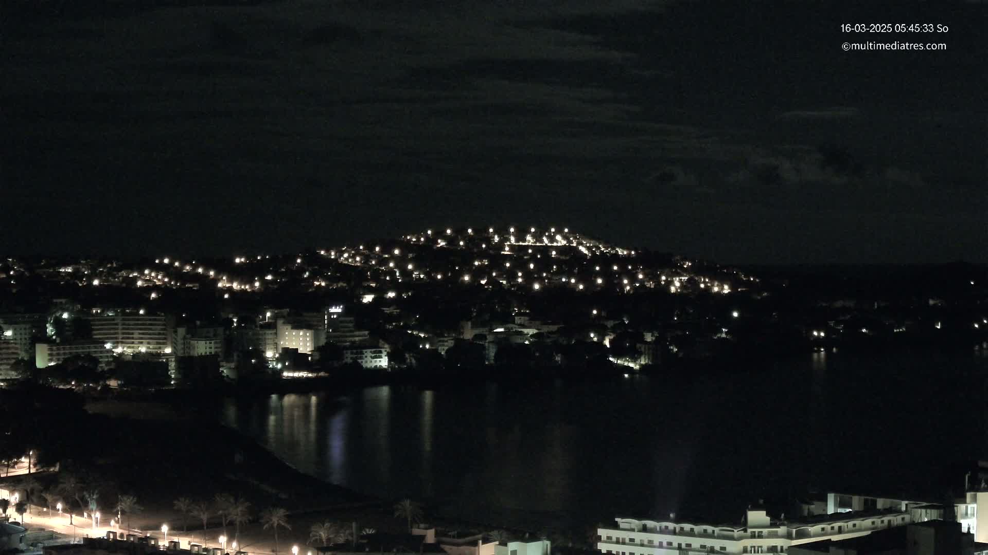 A nighttime view of a town situated on a hill overlooking a dark body of water under a mostly cloudy sky.