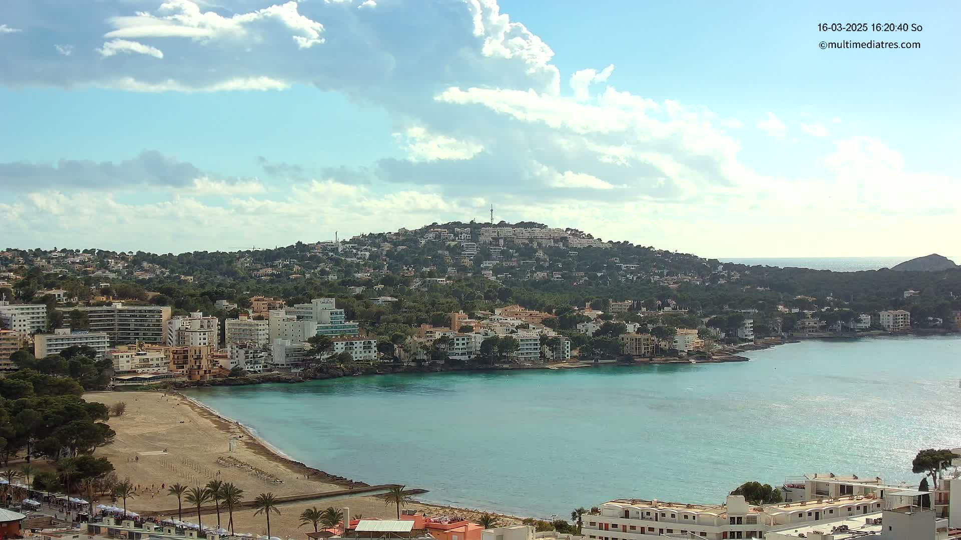 A coastal town nestled against a hillside, with a sandy beach bordering a calm, turquoise bay under a partly cloudy sky.