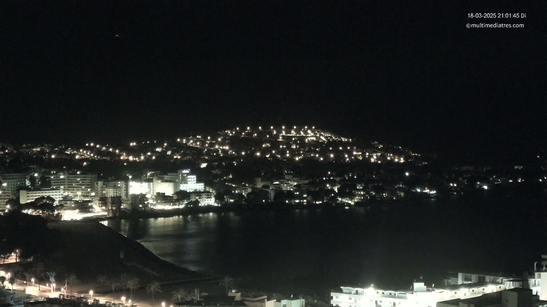 A nighttime view of a coastal town illuminated by many lights, situated on a hill overlooking a dark body of water under a clear sky.