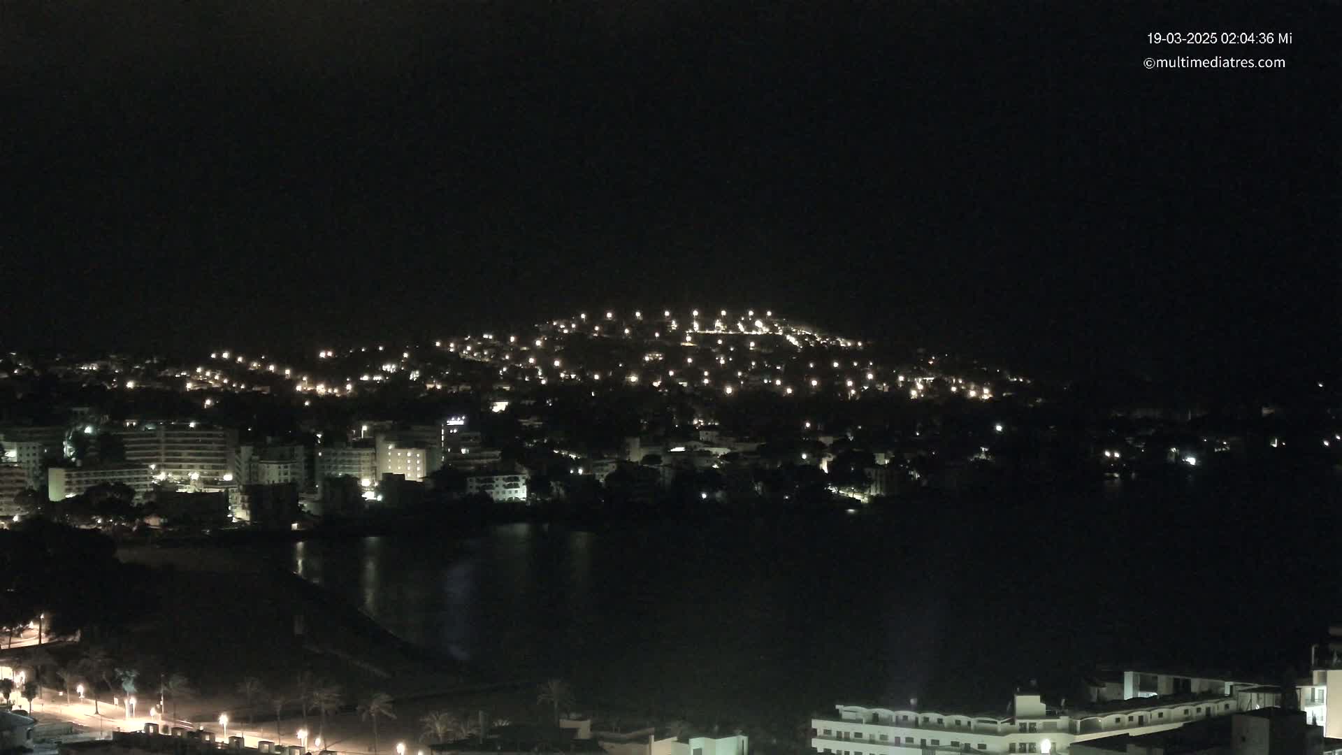 A nighttime view of a city illuminated by streetlights, situated alongside a dark body of water, with a hill dotted with lights in the background.