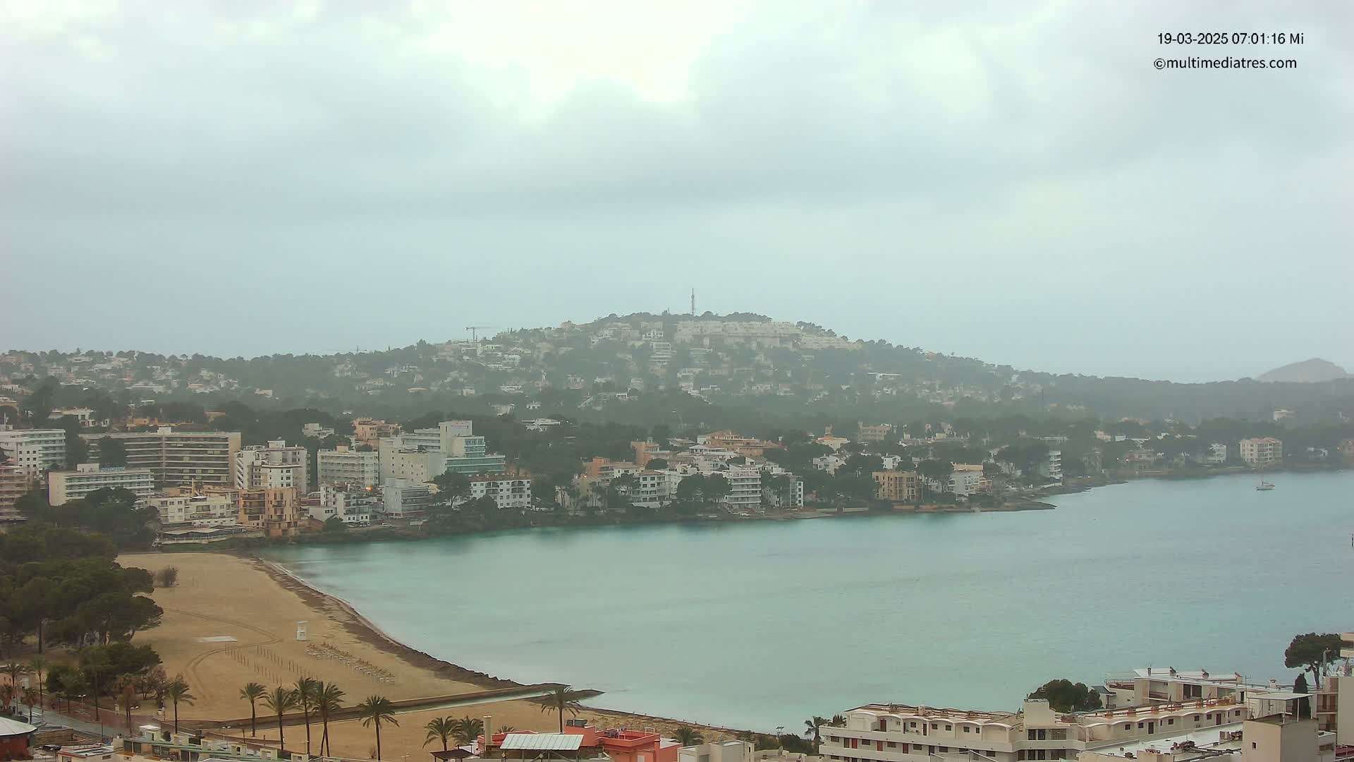 A hazy coastal town with a sandy beach and calm turquoise water is seen under an overcast sky.