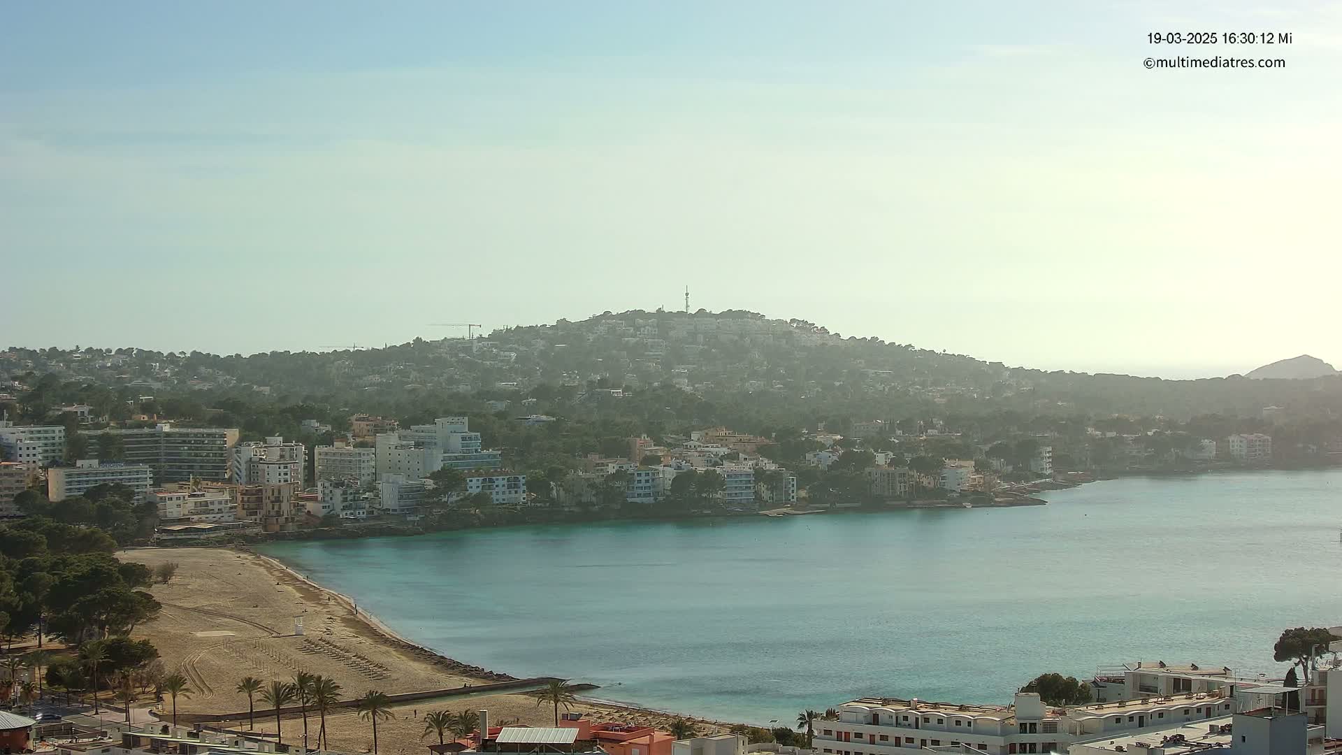 A hazy, sunny day overlooking a coastal town with a sandy beach, turquoise water, and buildings extending up a hillside.