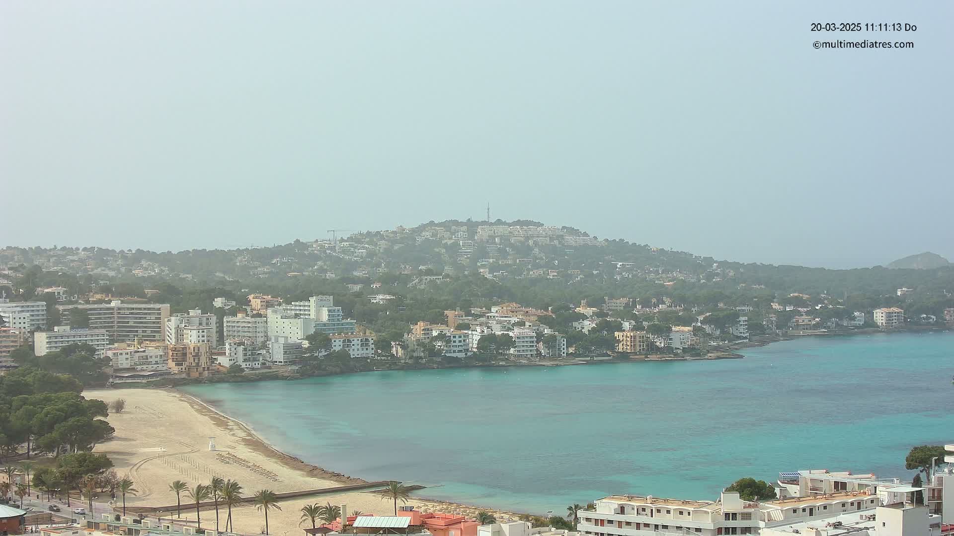 A hazy coastal town with turquoise water and sandy beach is nestled against a hill covered in buildings and trees.
