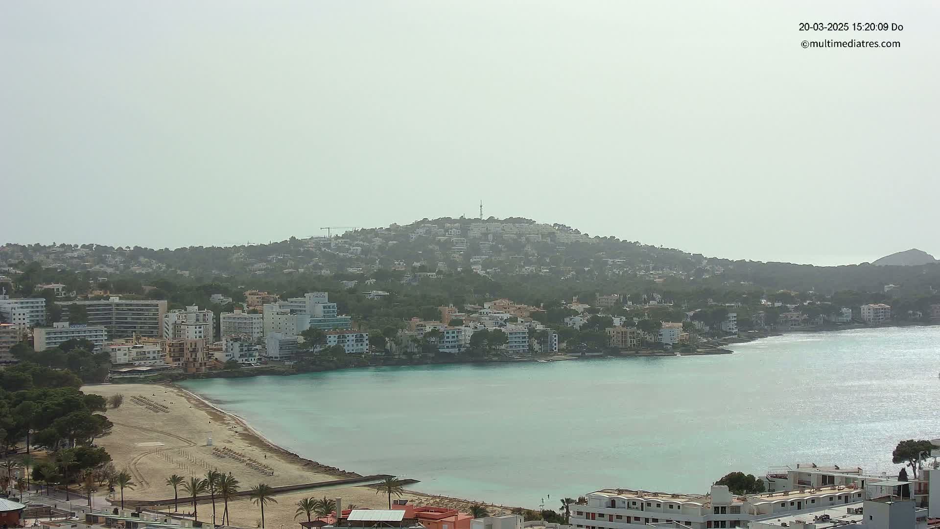 A hazy coastal town with buildings clustered around a calm, teal bay, backed by a hill covered in more buildings and vegetation.