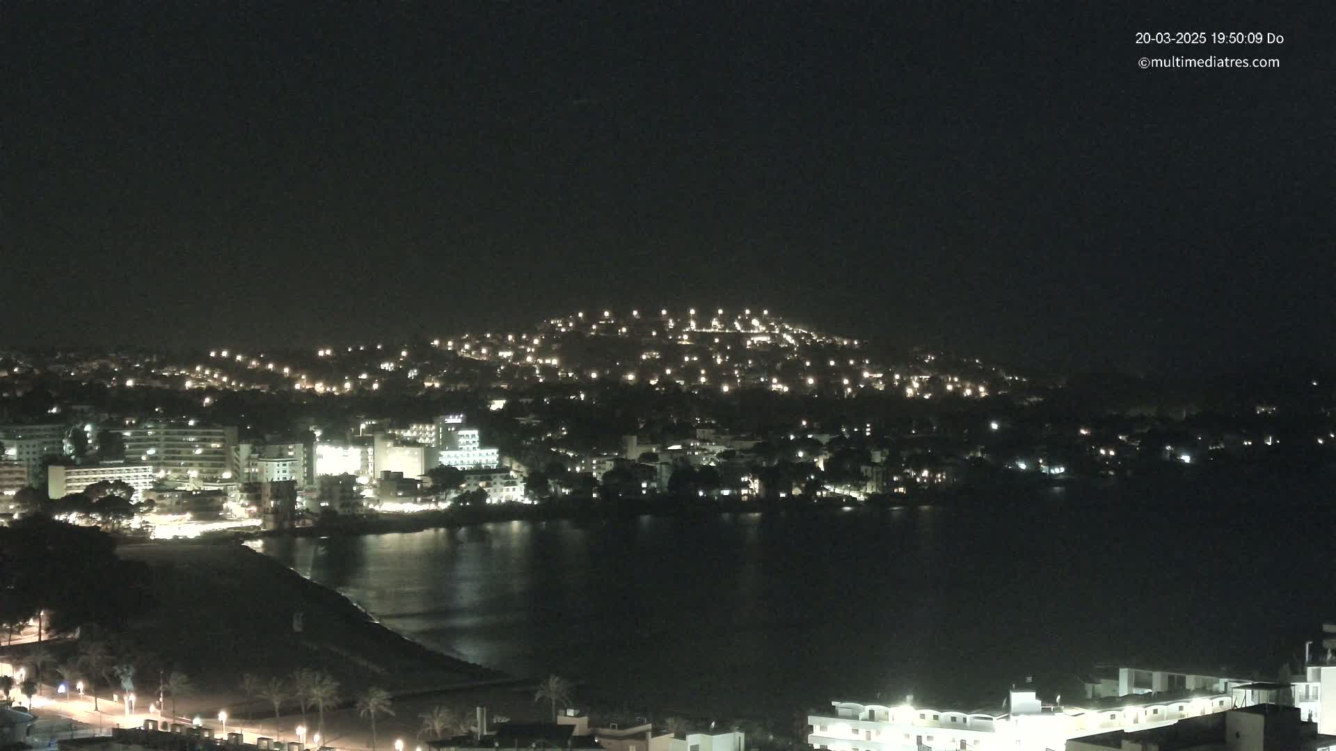 A nighttime view of a coastal town illuminated by lights, with a calm body of water in the foreground and a hill dotted with lights in the background under a clear, dark sky.