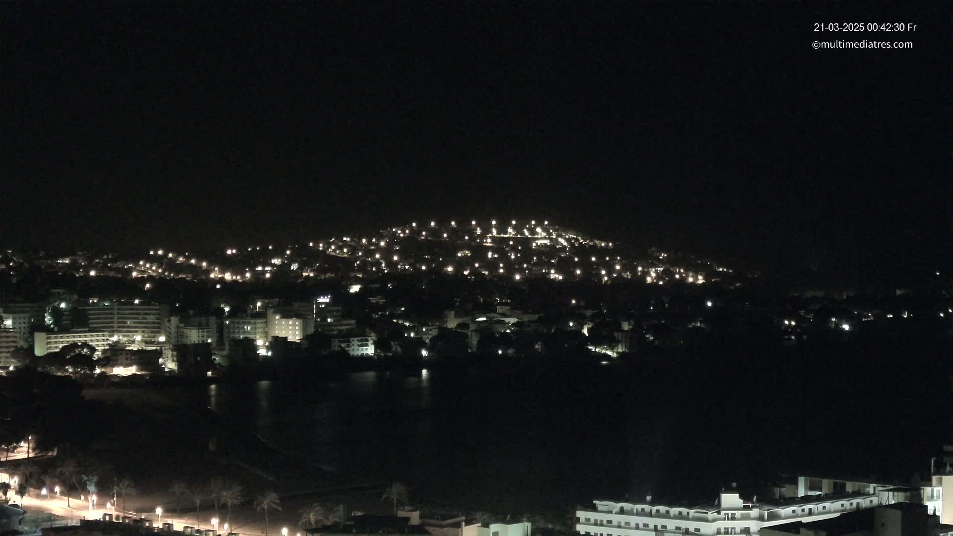 A nighttime view of a city illuminated by scattered lights, situated on a hill overlooking a dark body of water.