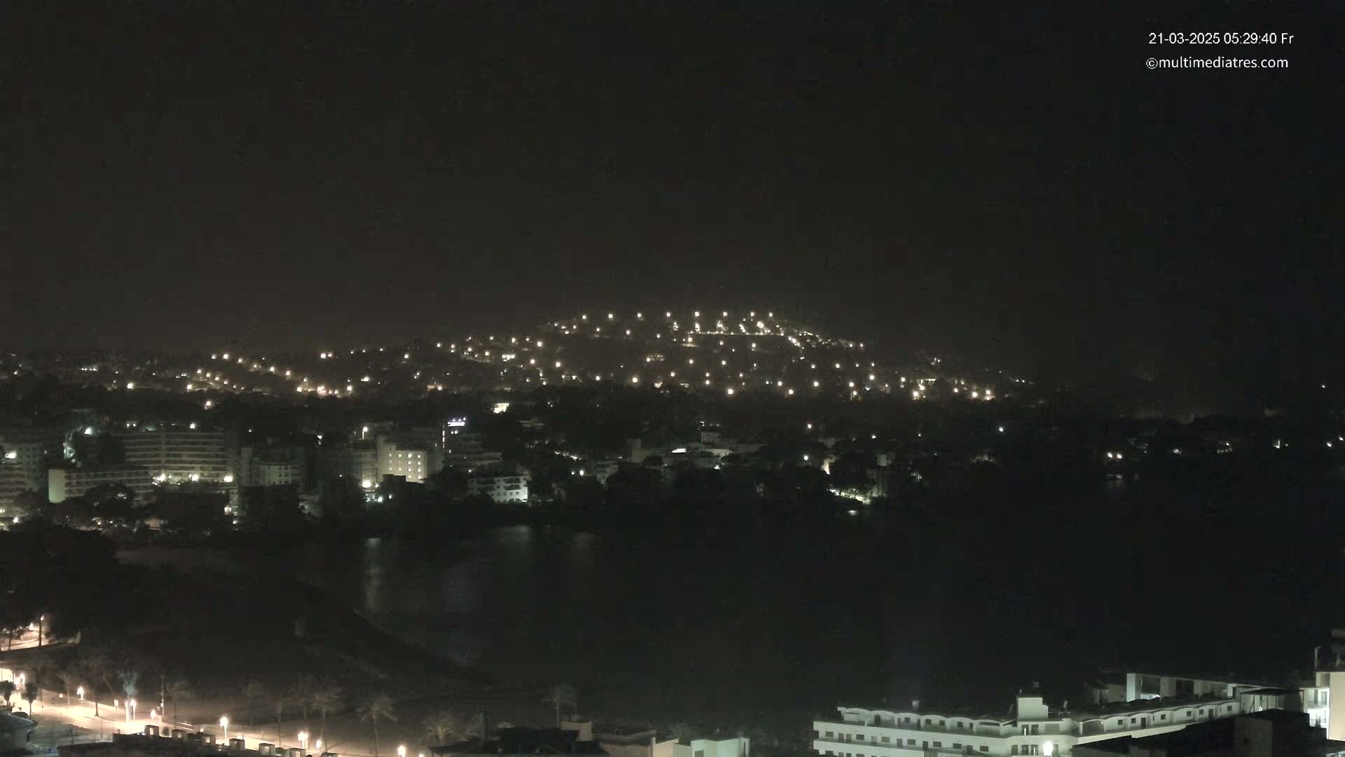 A nighttime view of a city illuminated by scattered lights, nestled against a dark hill and next to a body of dark water.