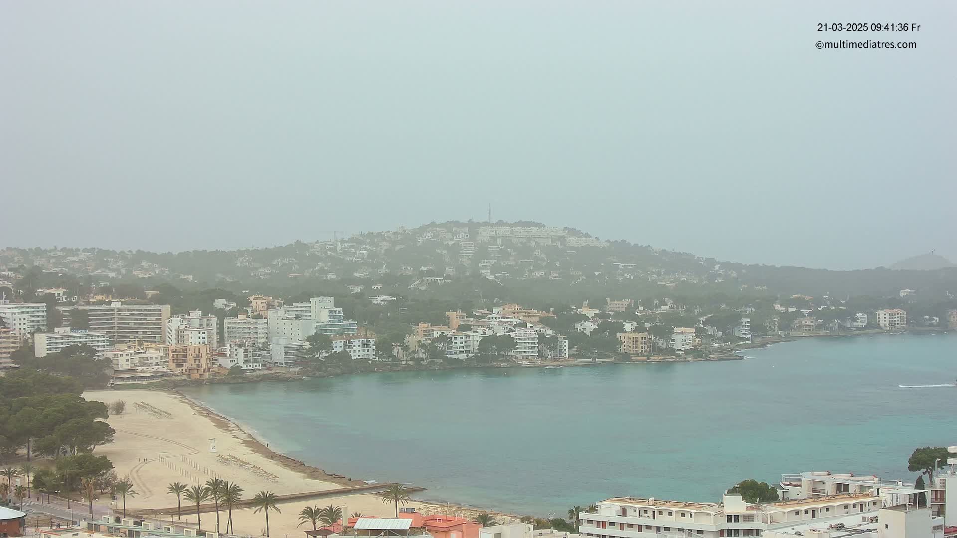 A hazy coastal town with a sandy beach and calm turquoise water is nestled below a hill covered in buildings and trees.
