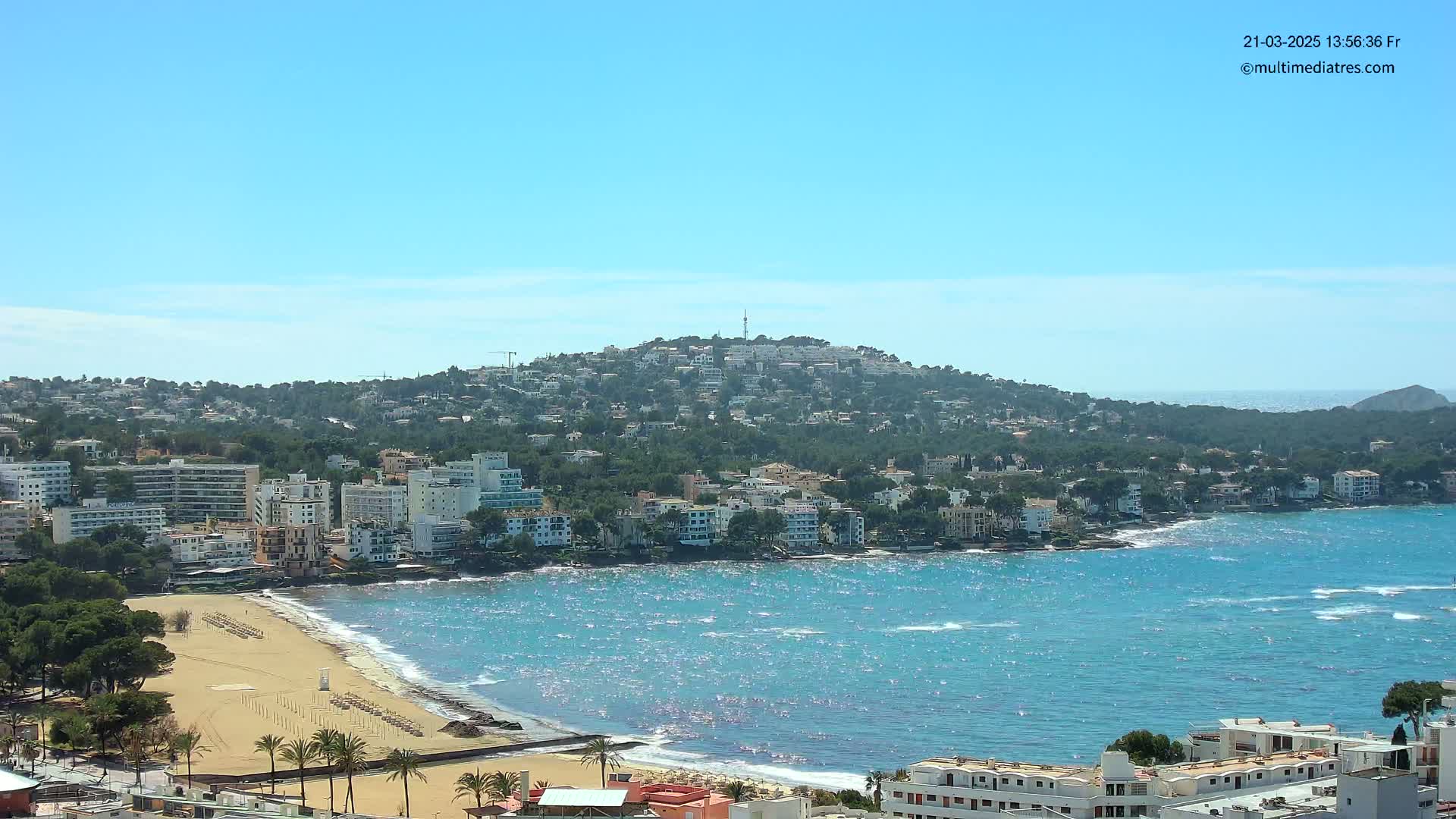 A sunny coastal town with a sandy beach and buildings that climb a hill overlooking a sparkling blue sea.
