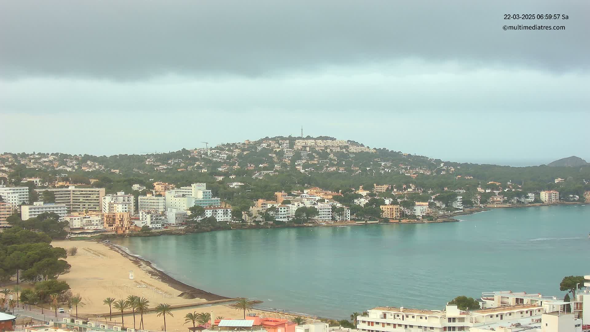 A coastal town with buildings clustered on a hillside and along a bay with a sandy beach, under an overcast sky.