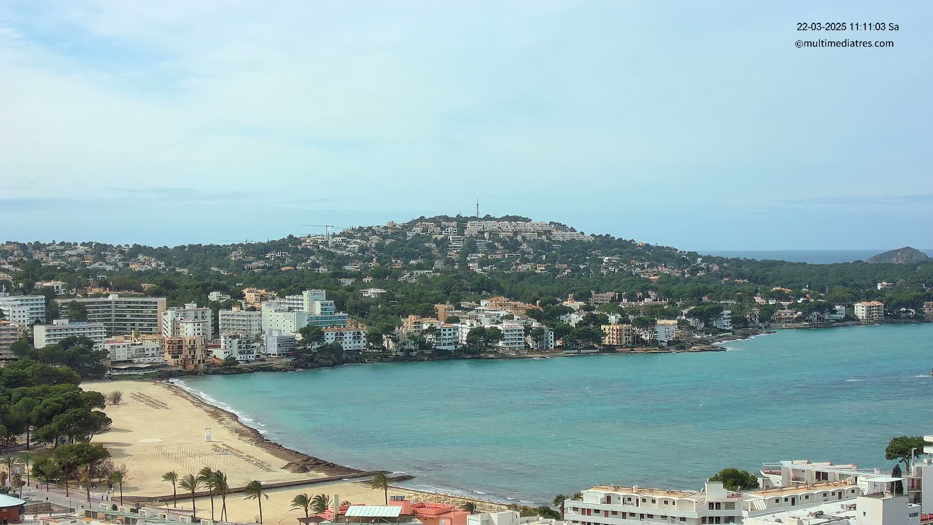 A coastal town with sandy beaches and buildings sits below a hill covered in houses and trees, under a mostly clear sky.