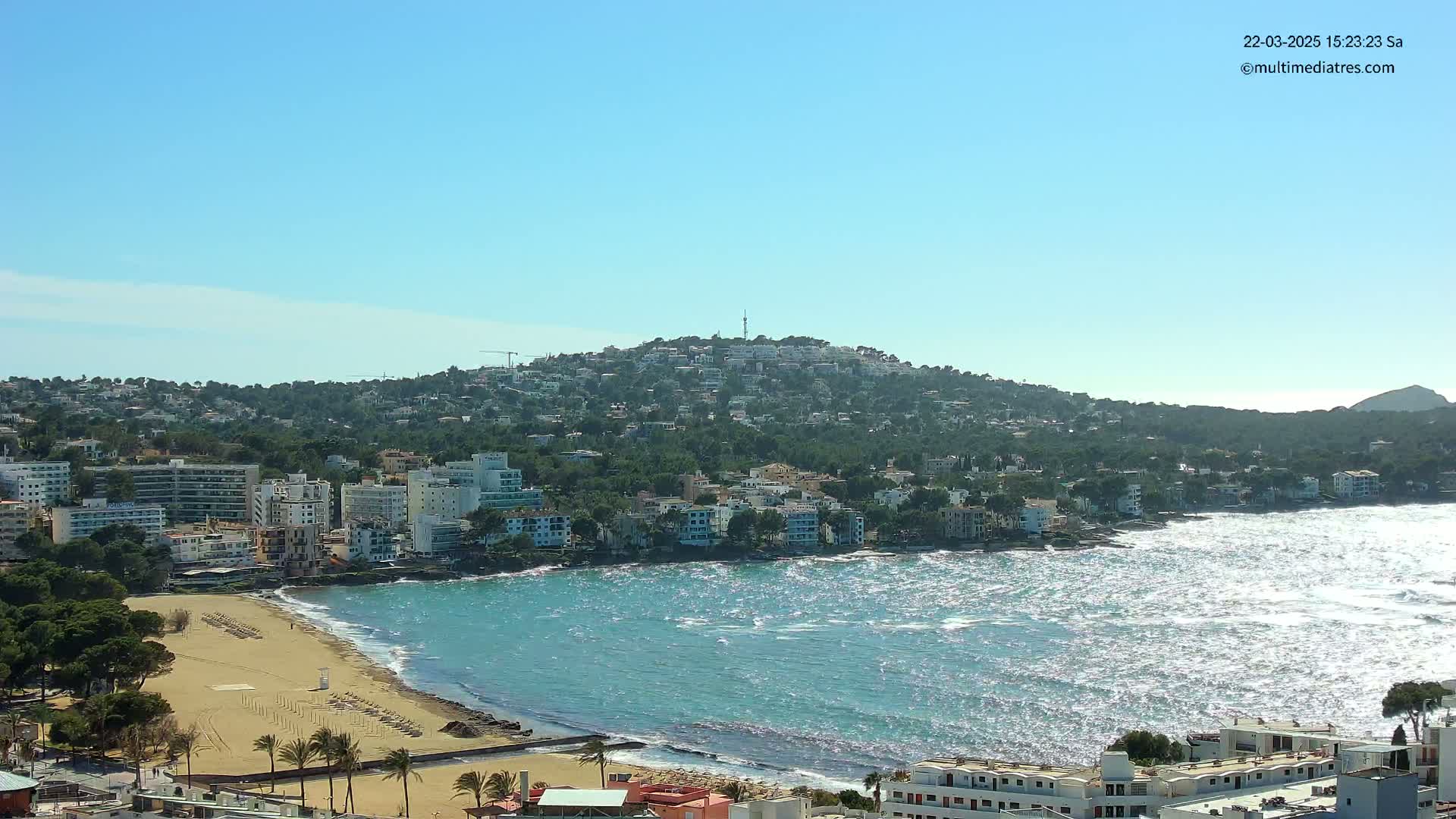 A coastal town with buildings on a hillside overlooking a sandy beach and a blue sea with choppy waves under a clear sky.