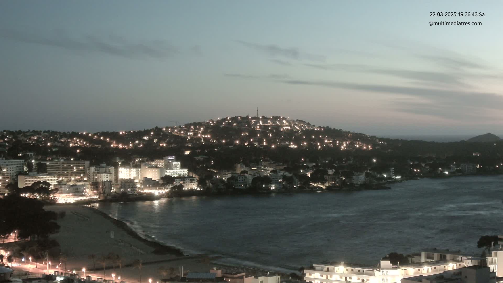 A coastal town at night, illuminated with lights, is situated on a gently sloping hill overlooking a dark bay with a calm sea.