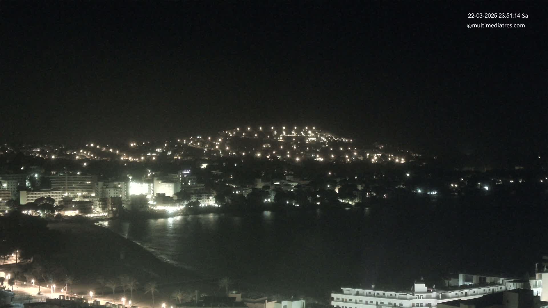 A nighttime view of a coastal town illuminated by lights, with a hill dotted with lights in the background and a dark body of water in the foreground.