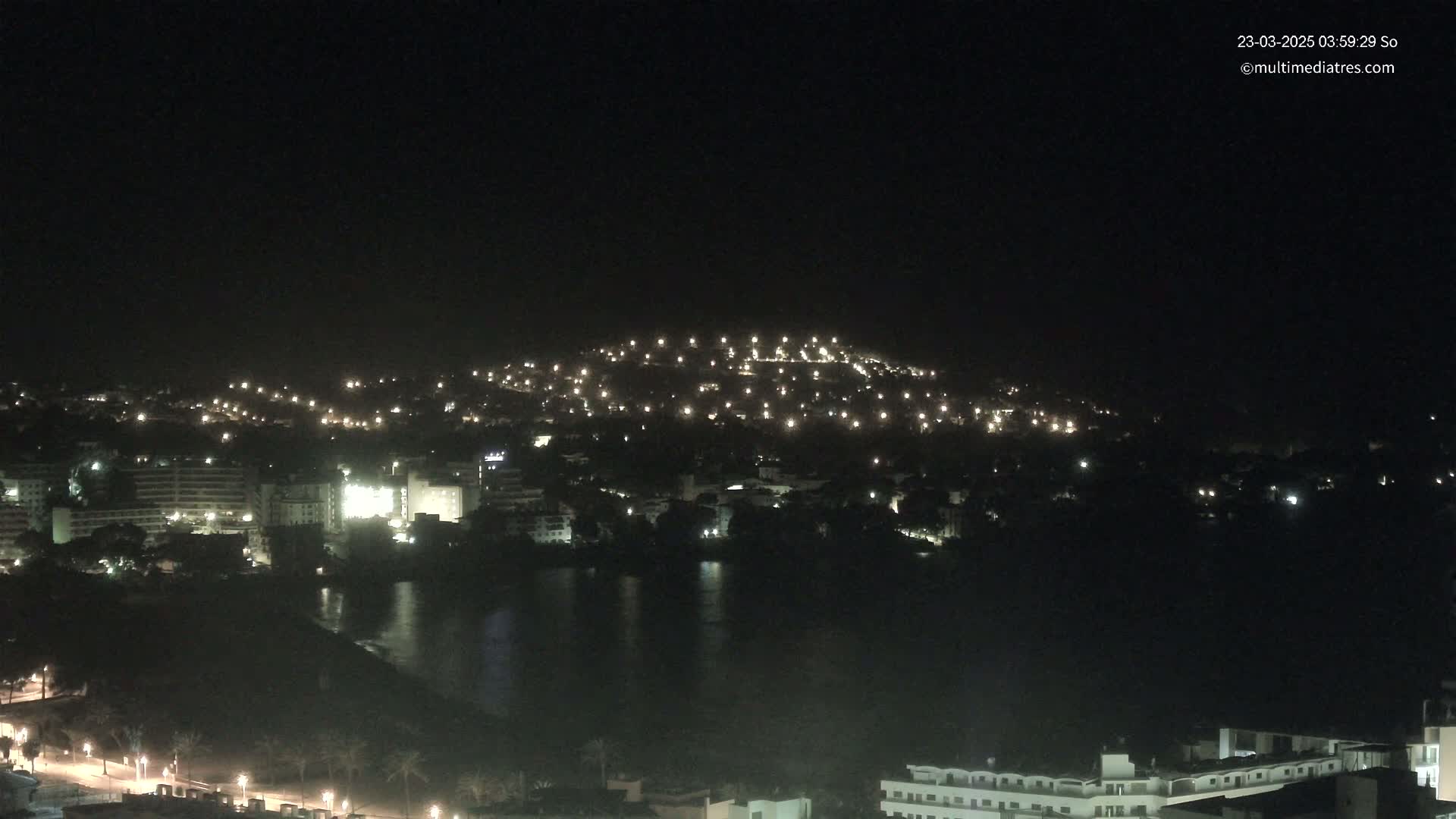 A nighttime view of a coastal town illuminated by lights, with a hill in the background under a clear, dark sky.