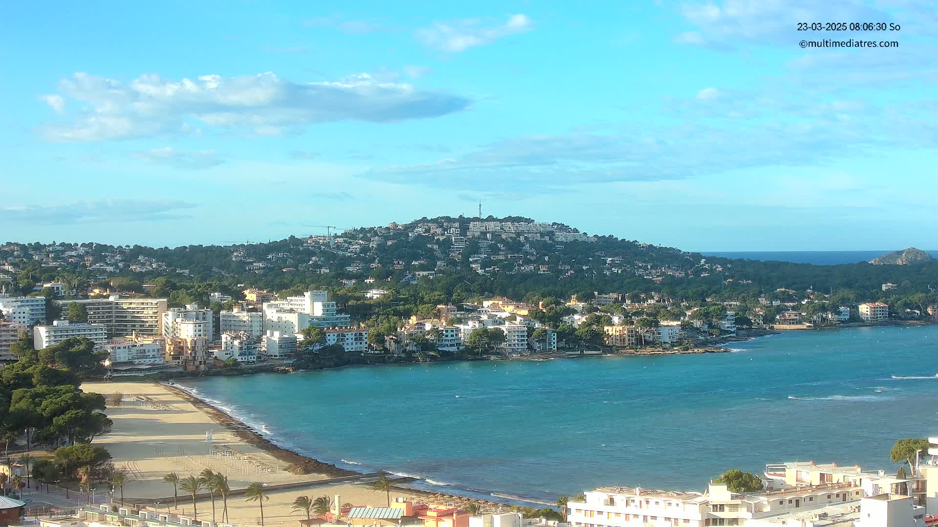 A coastal town with buildings on a hill overlooking a sandy beach and calm, turquoise water under a mostly clear sky.