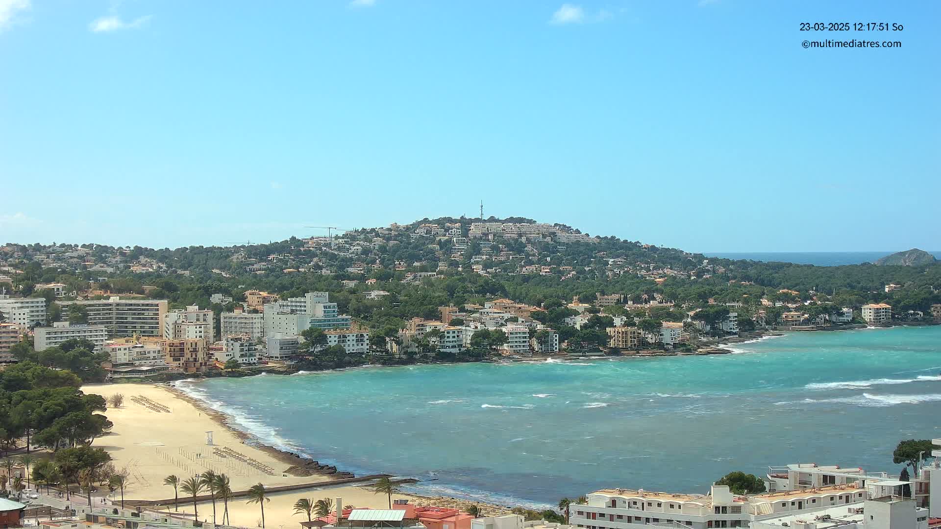 A coastal town nestled against a hill, with a sandy beach and turquoise water showing small waves under a clear blue sky.