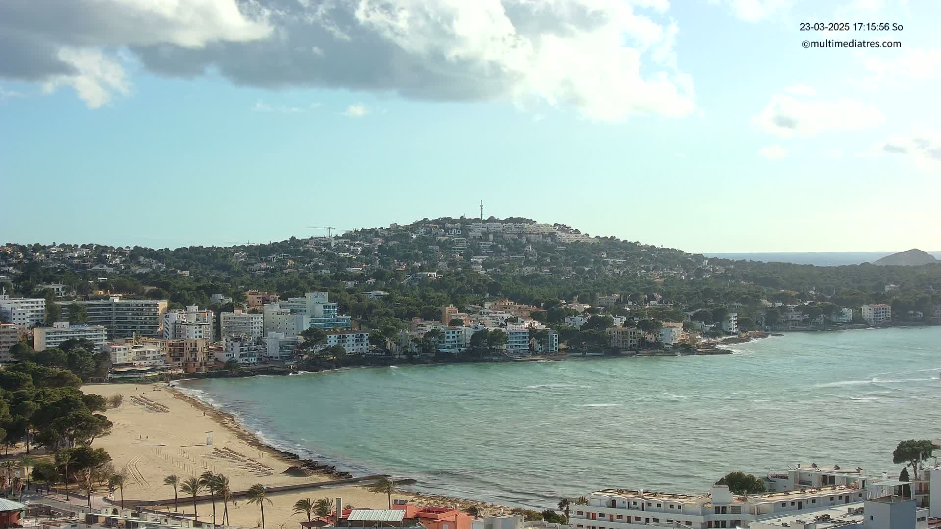 A partly cloudy day reveals a coastal town nestled against a green hill, with a sandy beach bordering a calm, teal sea.